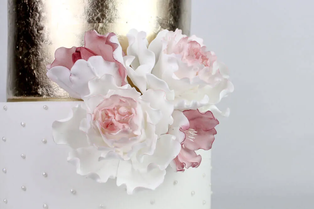A close-up of a decorated cake with white and pink sugar flowers affixed to its side. The cake has a white base with small round pearl-like details, and the top tier is covered in a shiny, gold metallic layer. The background is plain light gray.