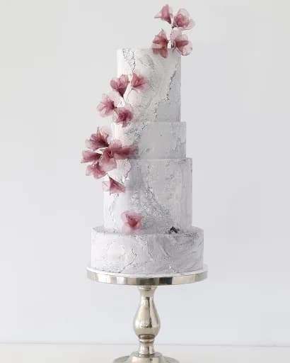 A four-tier white marble cake adorned with delicate pink floral decorations on each tier. The cake is elegantly positioned on a silver pedestal stand against a plain white background.