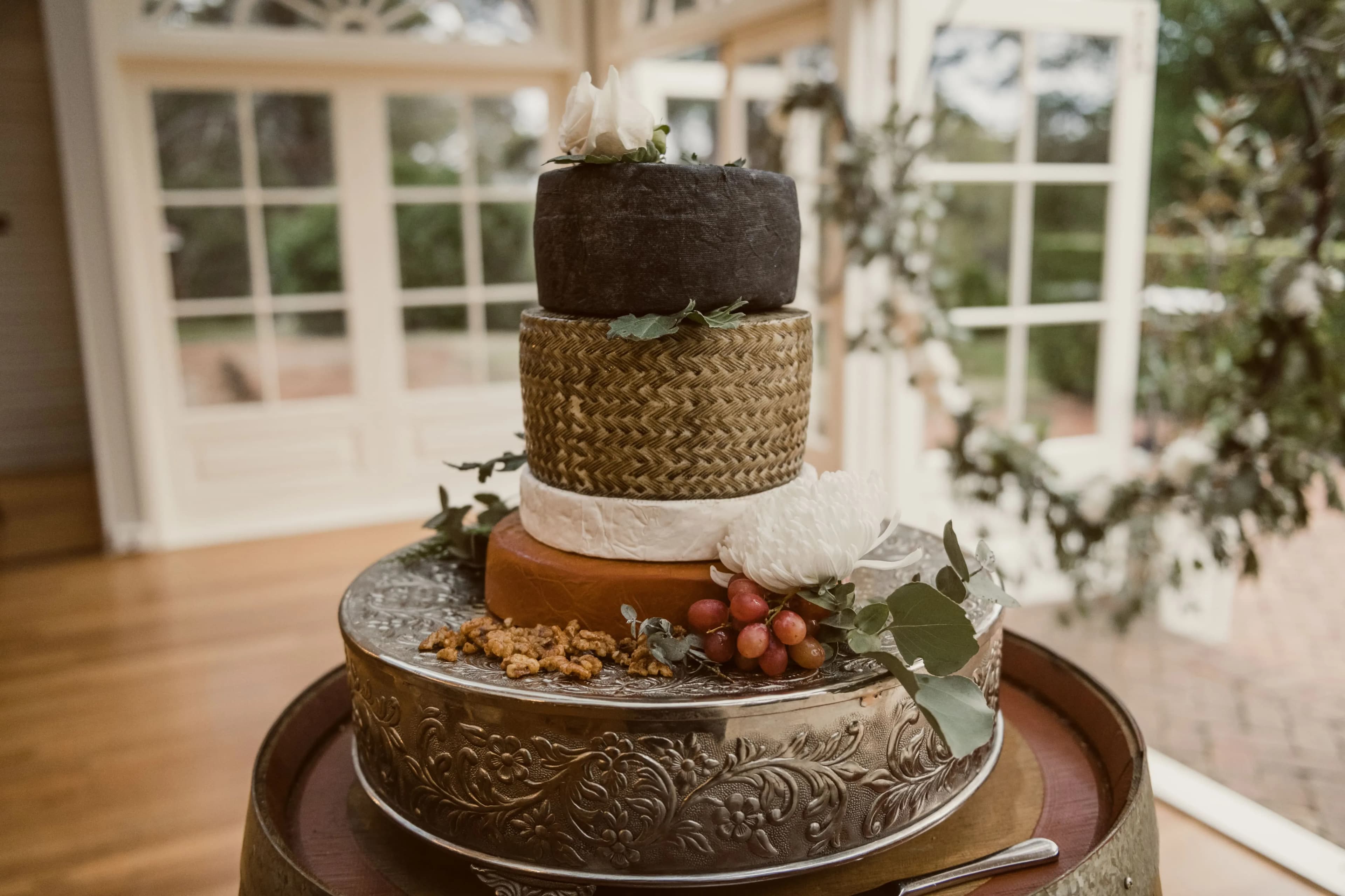 A four-tiered rustic wedding cake alternative made of stacked wheels of cheese with different colors and textures is decorated with grapes, nuts, and greenery. The cake is displayed on a silver tray atop a wooden barrel in a well-lit room with large windows.
