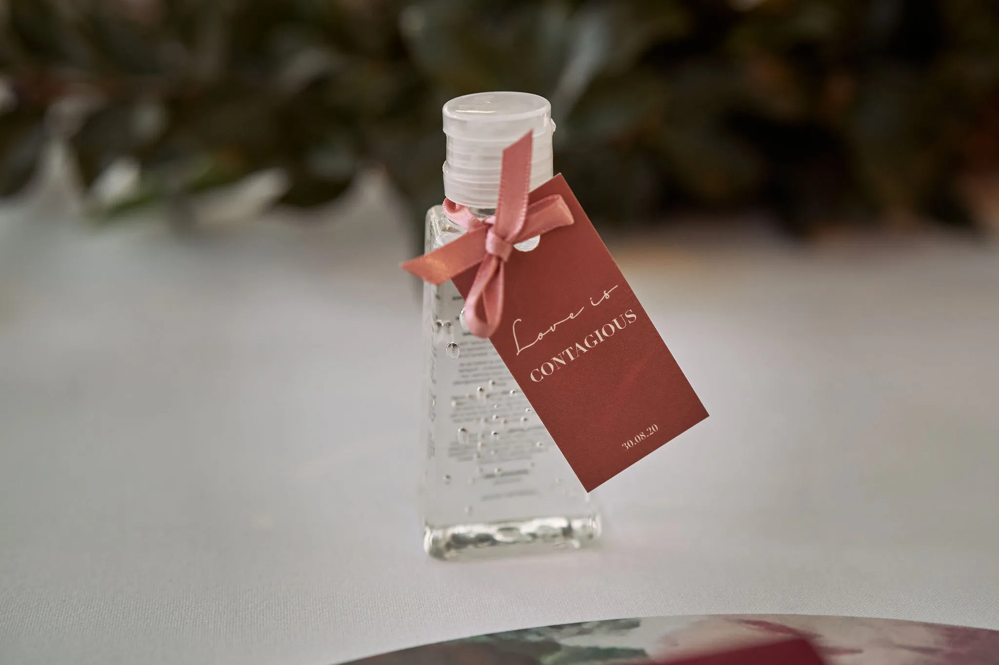 A small bottle of hand sanitizer with a clear gel inside is placed on a white surface. The bottle has a red tag tied with a pink ribbon that reads "Love is CONTAGIOUS." There are blurred green plants in the background.