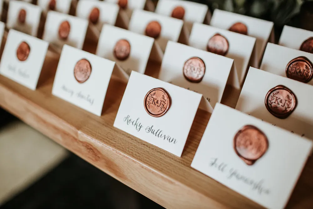 A display of white name cards with copper wax seals, arranged on a wooden surface. Each card is elegantly handwritten with different names, likely for an event or gathering. The wax seals feature intricate designs.