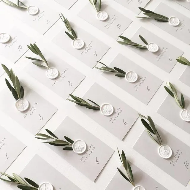 A grid of neatly arranged wedding place cards, each featuring a white wax seal with an initial, and adorned with a sprig of greenery lies on a white surface. The cards are numbered and list the names of guests in elegant black print.