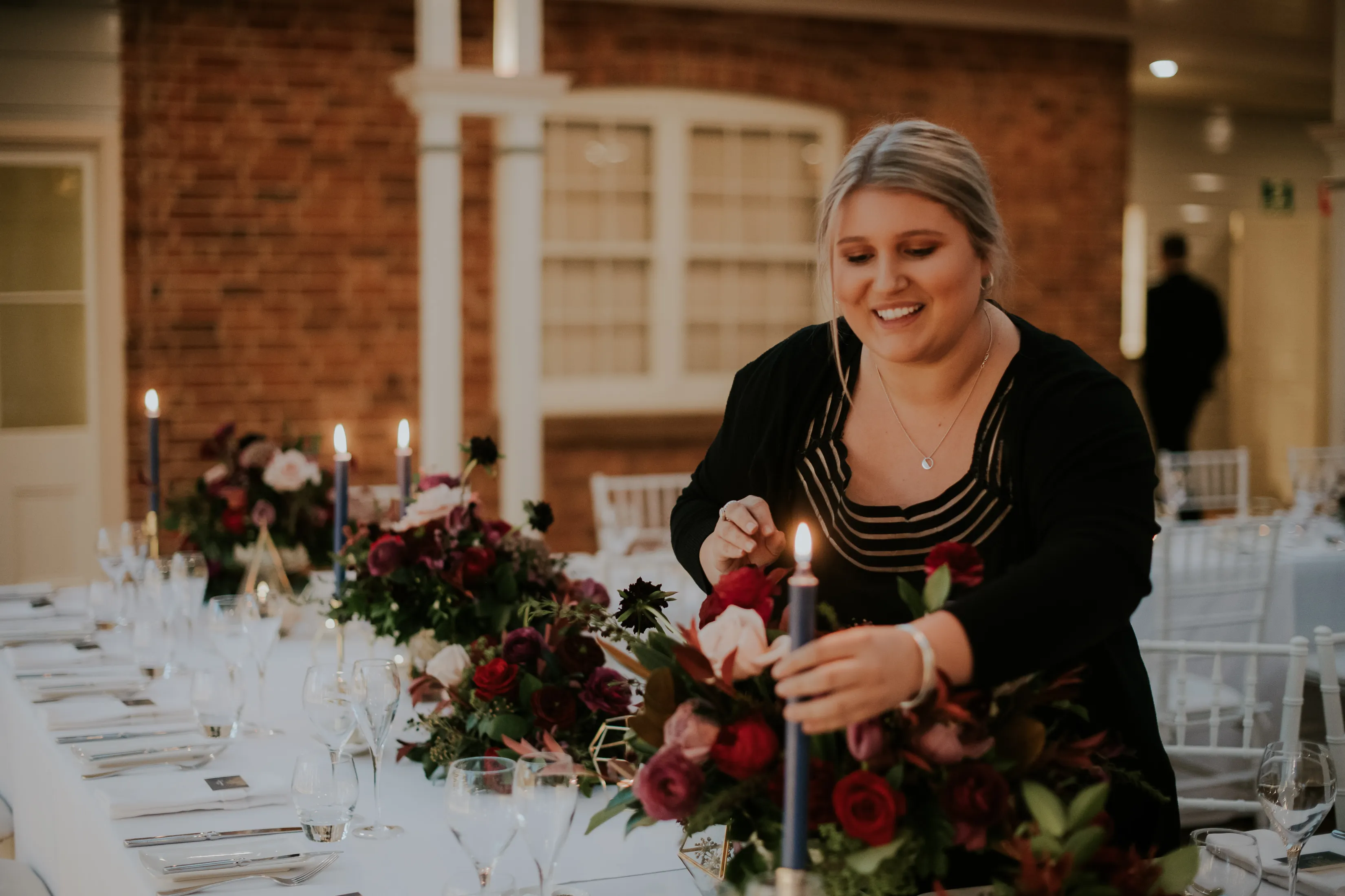 A woman with blonde hair is arranging a candle on a long dining table adorned with vibrant floral centerpieces and elegant glassware. She is smiling and appears to be preparing the table for an event in a well-lit room with a brick wall in the background.