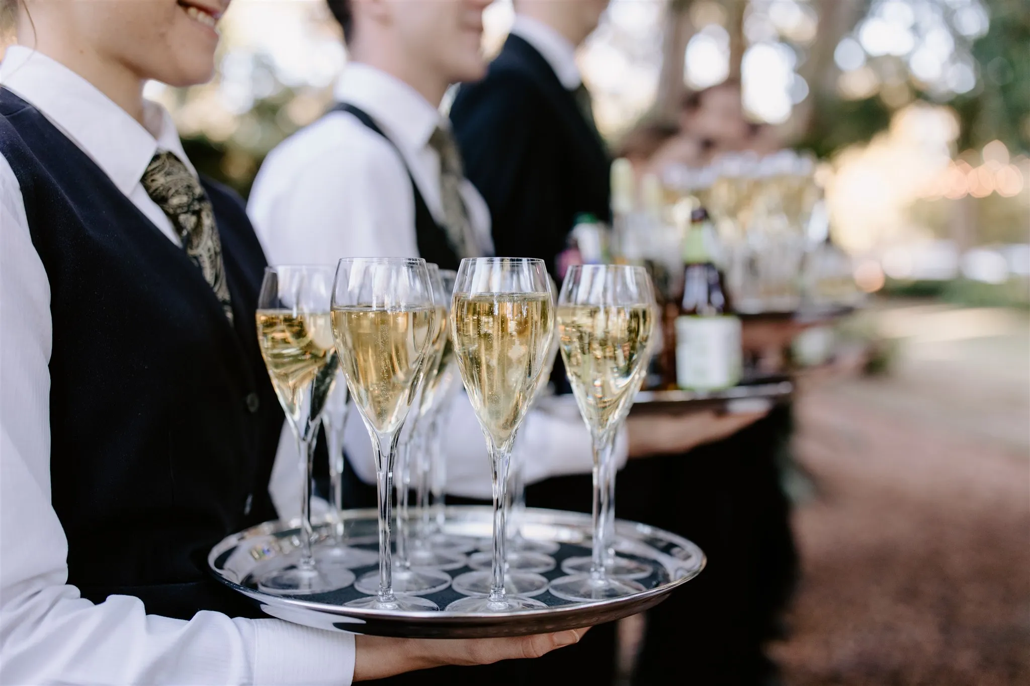 Servers carry trays of filled champagne flutes at an outdoor event. The servers are dressed in formal attire. The background shows blurry trees and lights indicating a festive atmosphere.