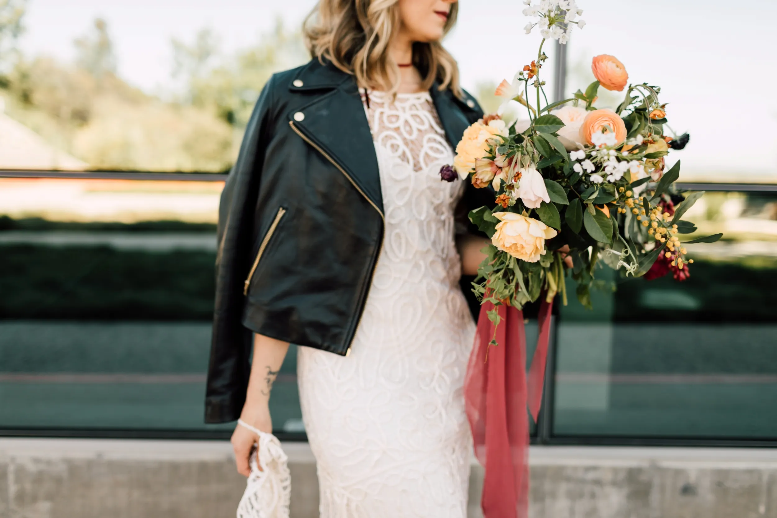 A woman in a white lace dress and black leather jacket stands outdoors, holding a bouquet of colorful flowers with orange, white, and green hues. She walks with a relaxed manner, her jacket draped over her shoulders, and carries a lace handkerchief in her right hand.