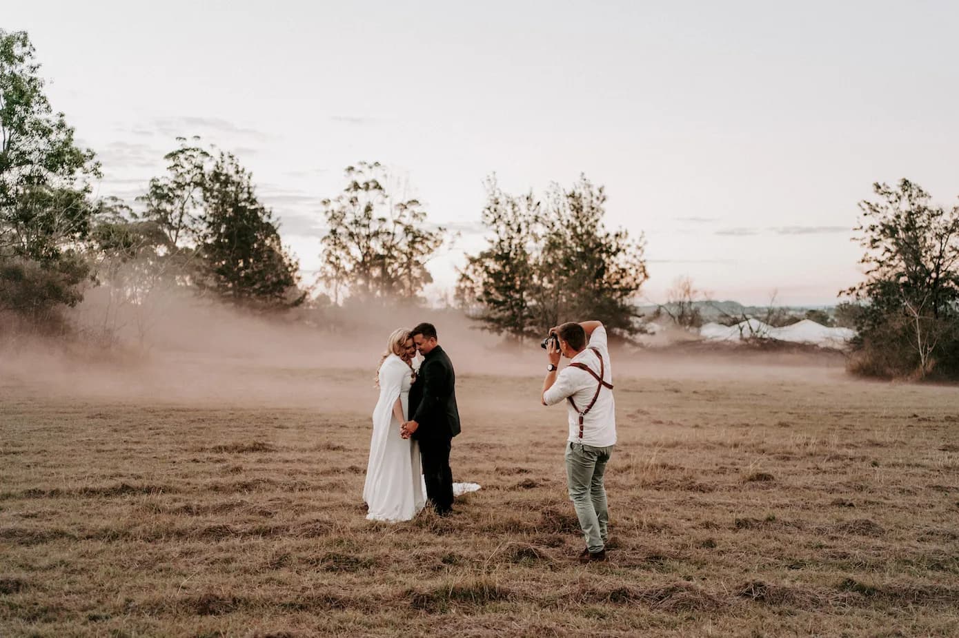 A couple in wedding attire embraces in an open field while a photographer captures the moment. The sky is at dusk, with light fog creating a dreamy atmosphere. Trees are in the background, and the couple gazes lovingly at each other.