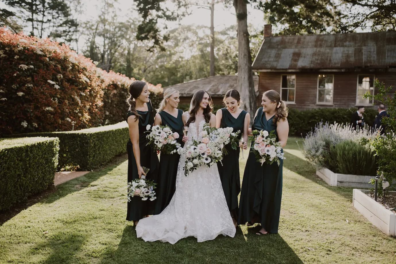 A bride in a white dress stands outdoors with four bridesmaids wearing dark green dresses. Each holds a bouquet of flowers. They are smiling and standing on a lawn with a rustic wooden house and manicured hedges in the background. The sun is shining.