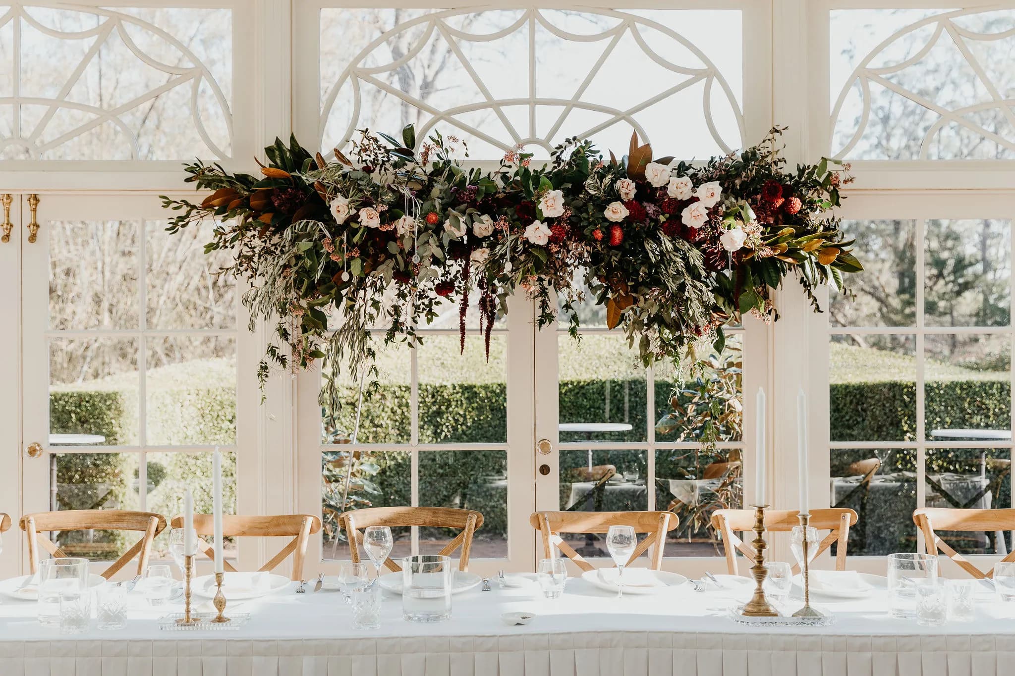 A beautifully decorated wedding reception table with a long, white tablecloth, elegant glassware, and gold candlesticks. Above the table is an elaborate floral arrangement featuring various flowers and greenery. Behind the table are large windows overlooking a garden.
.