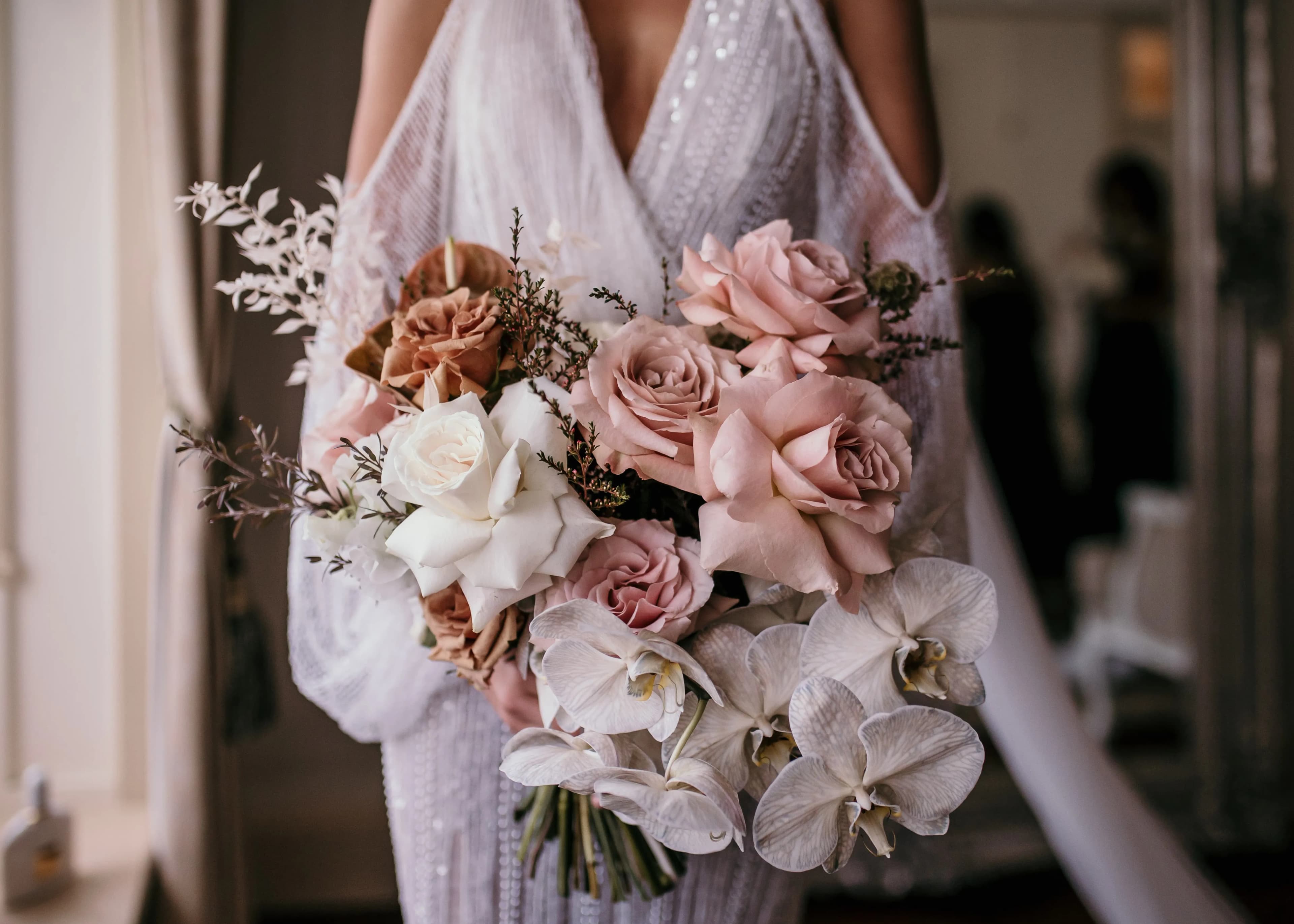 A bride holding a bouquet of flowers featuring a mix of white, pink, and peach roses, alongside white orchids. The bride is dressed in a detailed white gown with sheer sleeves. The background is softly lit, giving a warm and intimate ambiance.