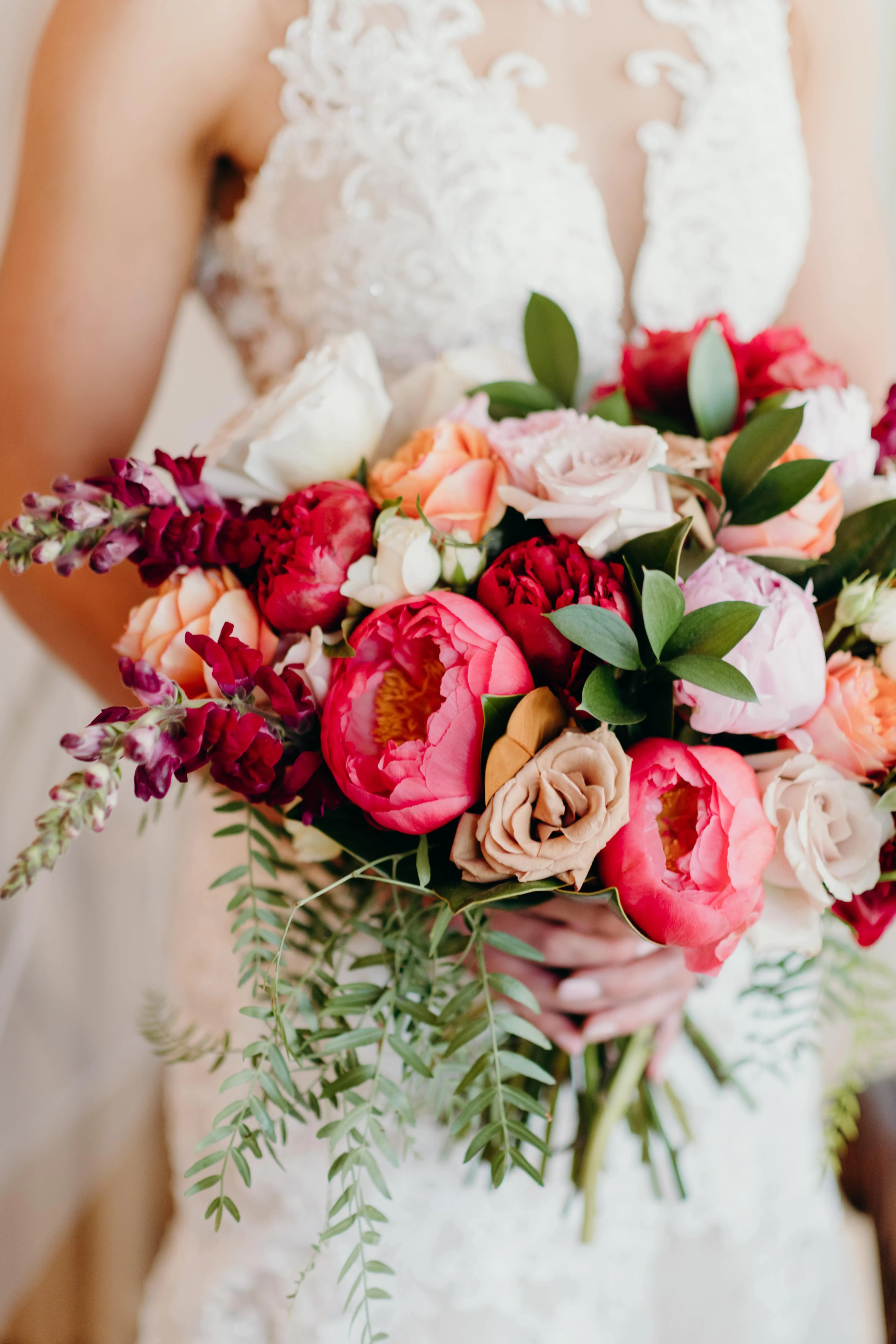 A bride in a white lace dress is holding a vibrant bouquet. The bouquet consists of pink peonies, red roses, peach and white flowers, and various green leaves. The intricate floral arrangement contrasts beautifully with the delicate lace of her gown.