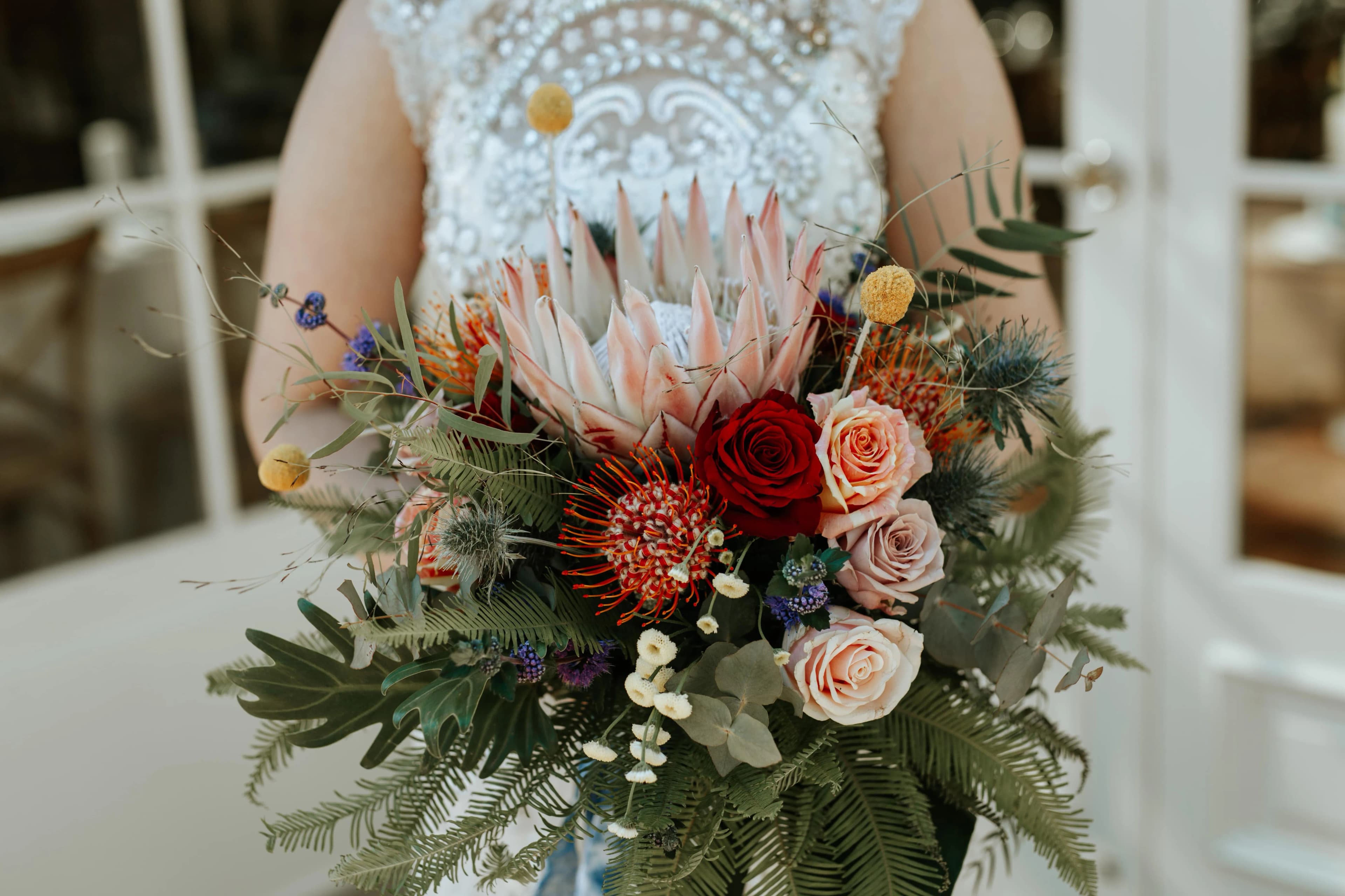 A person in a detailed white lace outfit is holding a bouquet featuring a large pink protea flower, red and blush roses, yellow billy balls, and various greenery and filler flowers. The background shows a bright interior with glass doors.