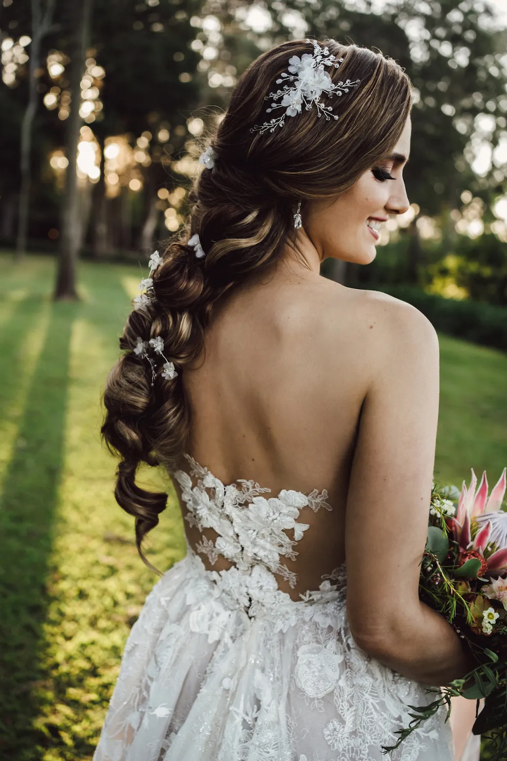 A bride stands outdoors in a sunlit garden, looking over her shoulder with a smile. She is wearing a white lace dress with floral details and a long braid adorned with hairpieces. She holds a bouquet of flowers and the sunlight filters through the trees behind her.