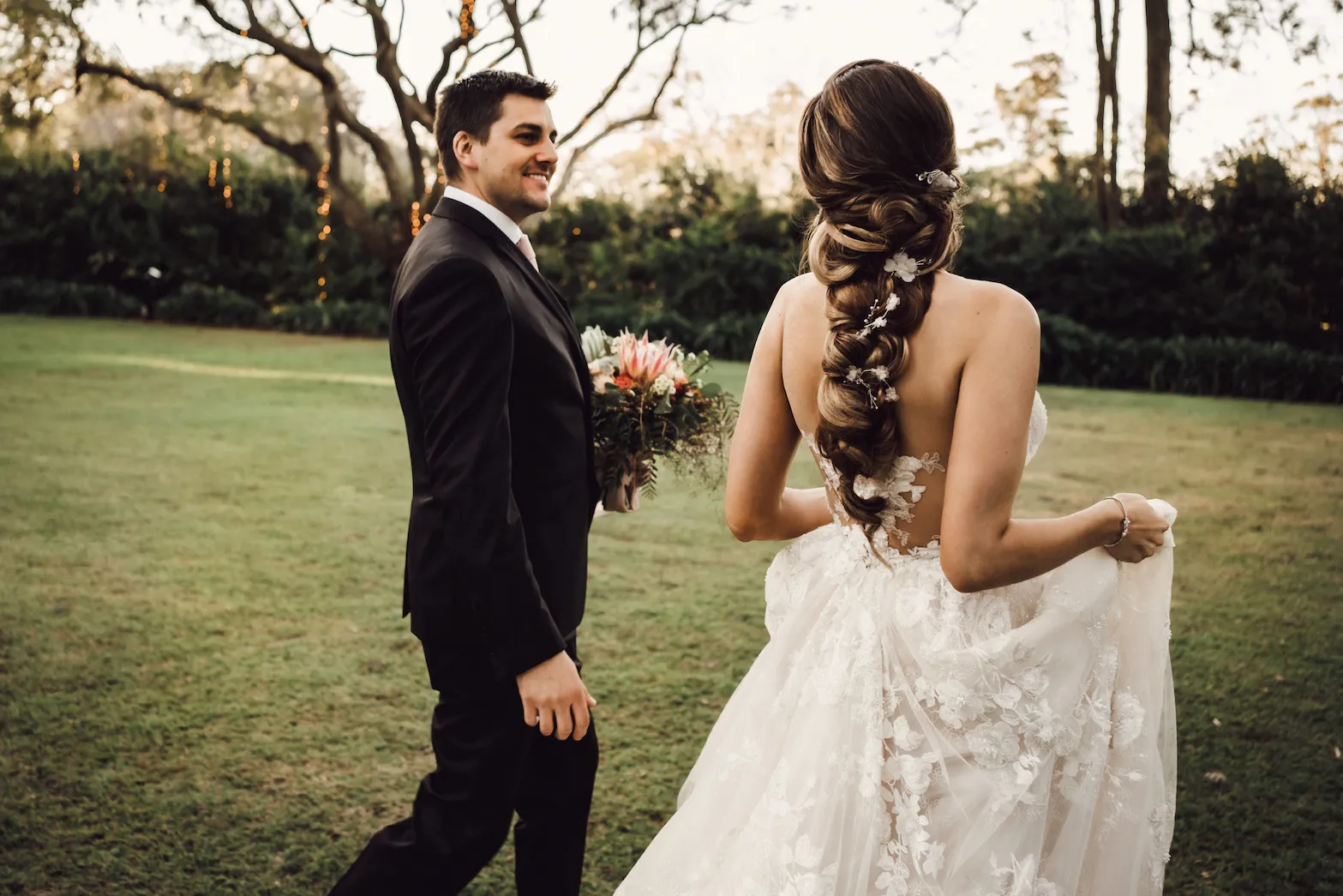 A bride and groom walk hand in hand on a grassy lawn. The bride's long, braided hair is adorned with flowers, and she wears a flowing, white, lace wedding dress. The groom is in a dark suit and holds a bouquet. Trees with soft lighting are in the background.