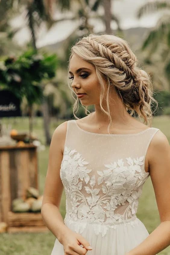 A bride with intricate braided blonde hair and a neutral makeup look stands outdoors. She wears a sleeveless white gown with a sheer neckline and floral lace details. The background features blurred greenery and a wooden structure set for an outdoor event.