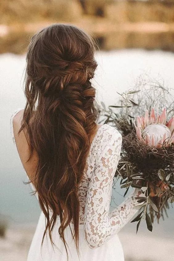 A woman with long, wavy brunette hair styled in an elaborate braid stands with her back to the camera. She wears a white lace dress and holds a bouquet featuring a large, pale pink King Protea flower. The background is blurred, highlighting a body of water and greenery.