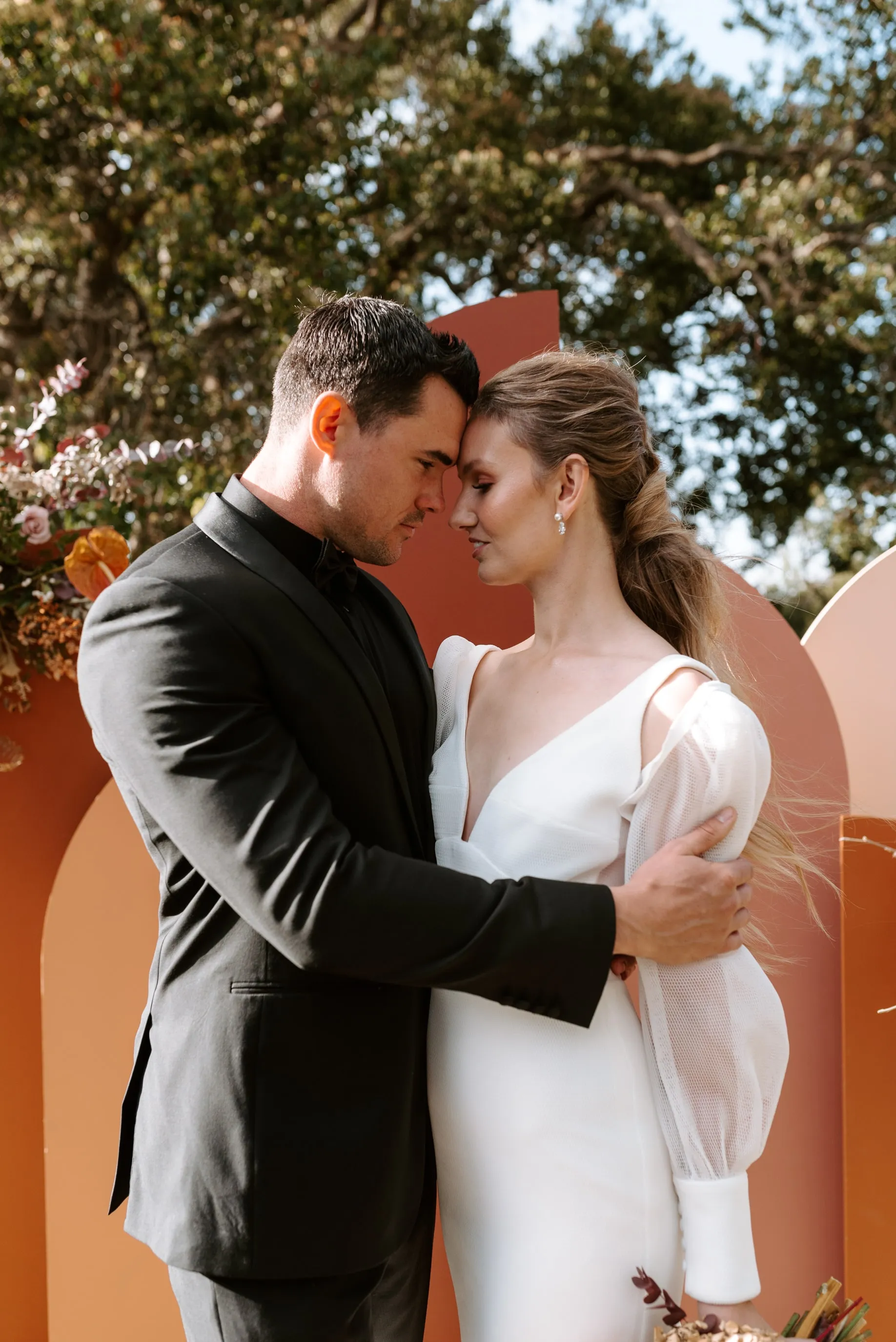 A couple dressed in formal attire embraces lovingly outdoors. The woman wears a white dress with sheer sleeves, and the man wears a black suit. They stand in front of a decorative backdrop with flowers, trees, and wooden arches. Their foreheads touch gently.