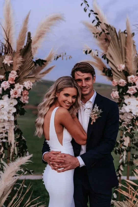 A bride and groom embrace each other and smile in front of a wedding arch decorated with pampas grass and roses. The bride is wearing a white dress with an open back, while the groom is in a dark suit with a boutonniere. The background shows a scenic outdoor setting.