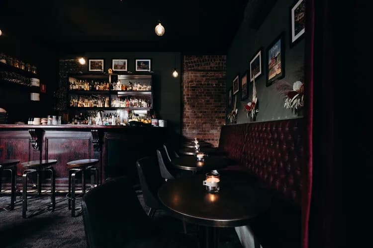 A dimly lit bar features a wooden bar counter with bar stools and a well-stocked shelf of bottles. Alongside is a line of small round tables with red cushioned seating. Framed pictures adorn the dark walls, and small lit candles create a cozy ambiance.