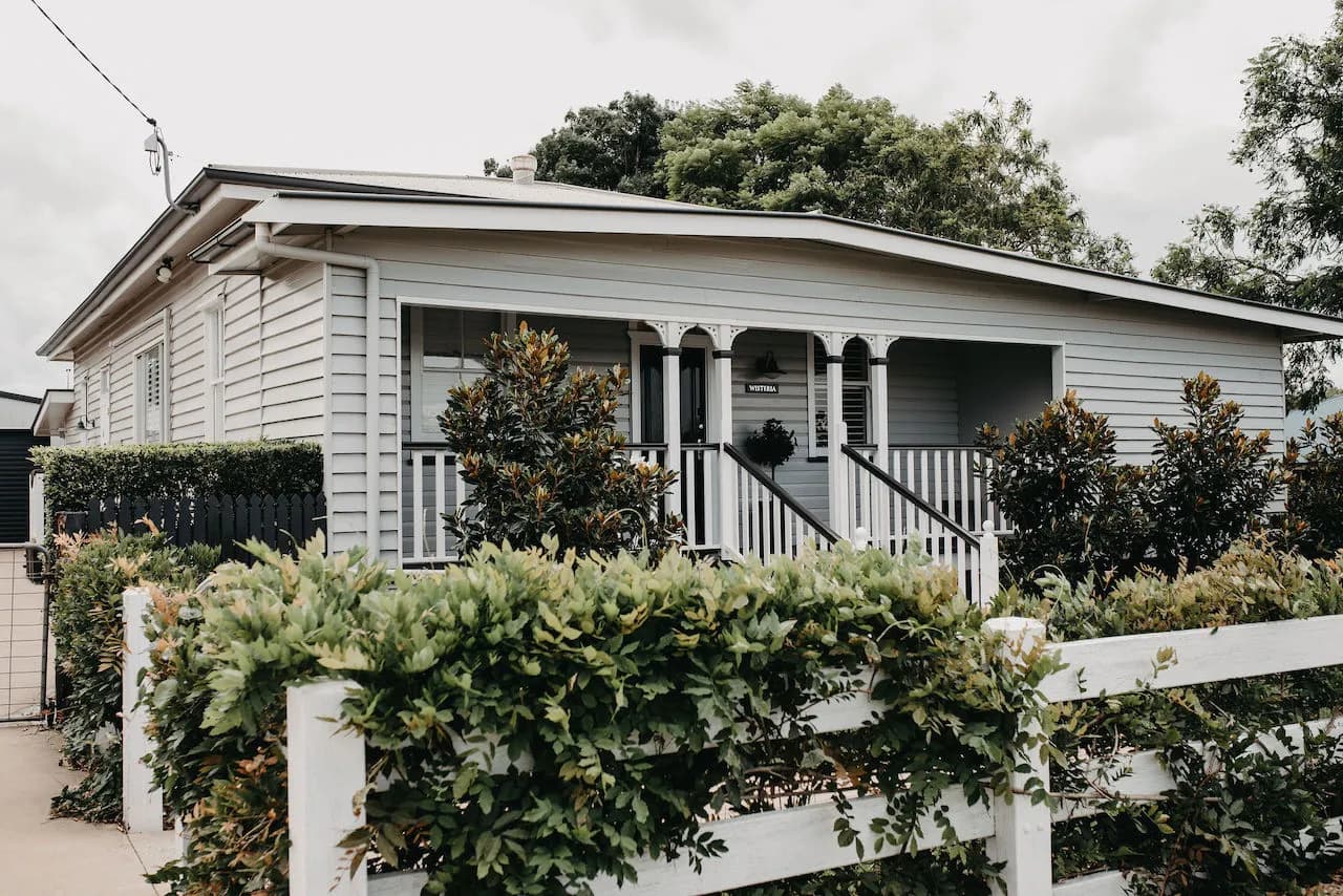 A cozy, single-story white house with a decorative porch and railings is surrounded by lush greenery and bushes. The grass is neatly mowed, and a white picket fence borders the front yard. The sky above is overcast with light gray clouds.