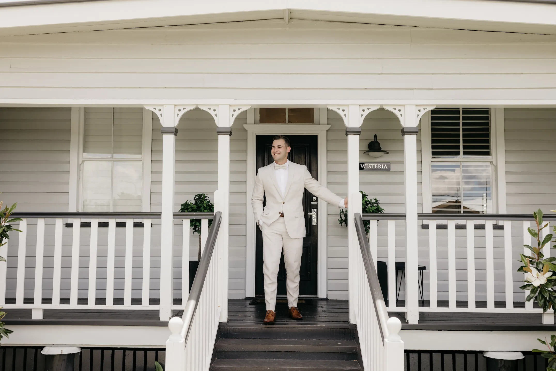 A man in a light-colored suit stands on the porch of a white house with a sign reading "Wisteria." He is smiling, with one hand resting on a railing, and the house has black steps, white railings, and two potted plants flanking the entrance.