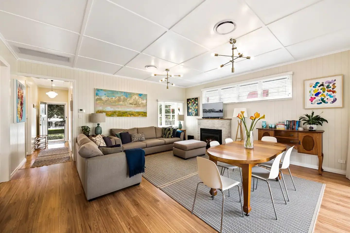 A modern, open-plan living and dining area featuring neutral-toned furniture. The room includes a gray sectional sofa with a coffee table, a round wooden dining table with white chairs, colorful wall art, a fireplace, and large windows letting in natural light.