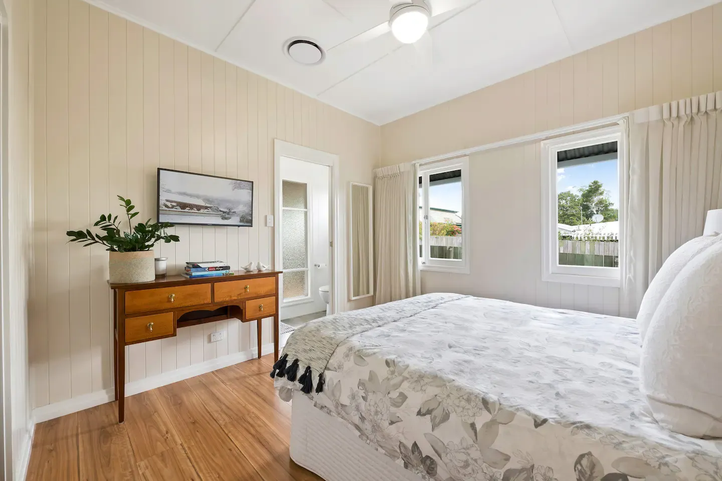 A cozy bedroom with light-colored wood paneling on the walls and hardwood flooring. It features a neatly made bed with floral bedding, a wooden dresser with plants and books, a ceiling fan, and large windows with white curtains allowing natural light in. A doorway leads to an adjoining bathroom.