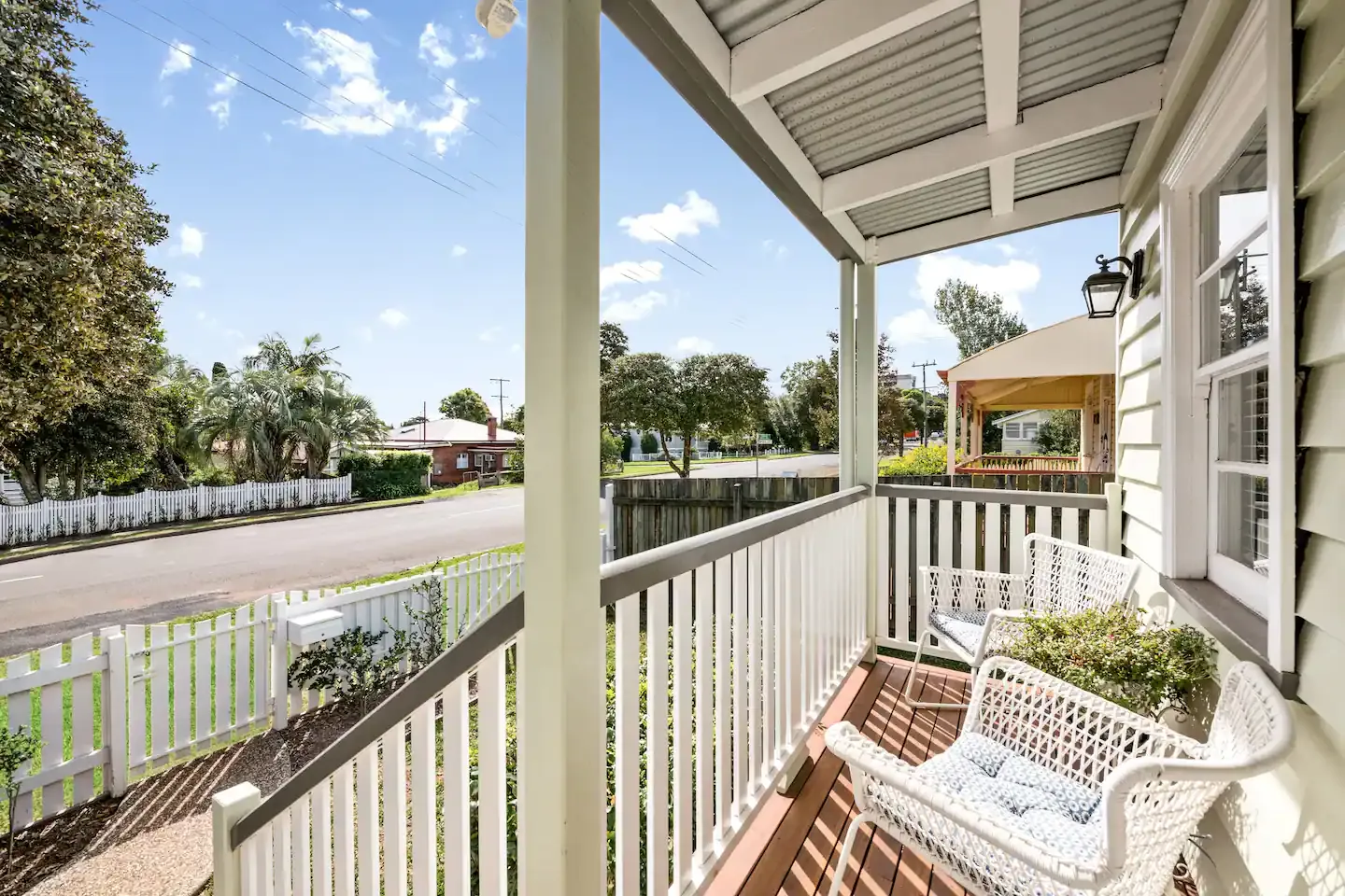 The image shows a cozy front porch with white wooden railings, wicker chairs, and a view of a quiet suburban street lined with trees and houses. The porch provides a sunny spot for relaxation, facing a well-maintained lawn and garden area.