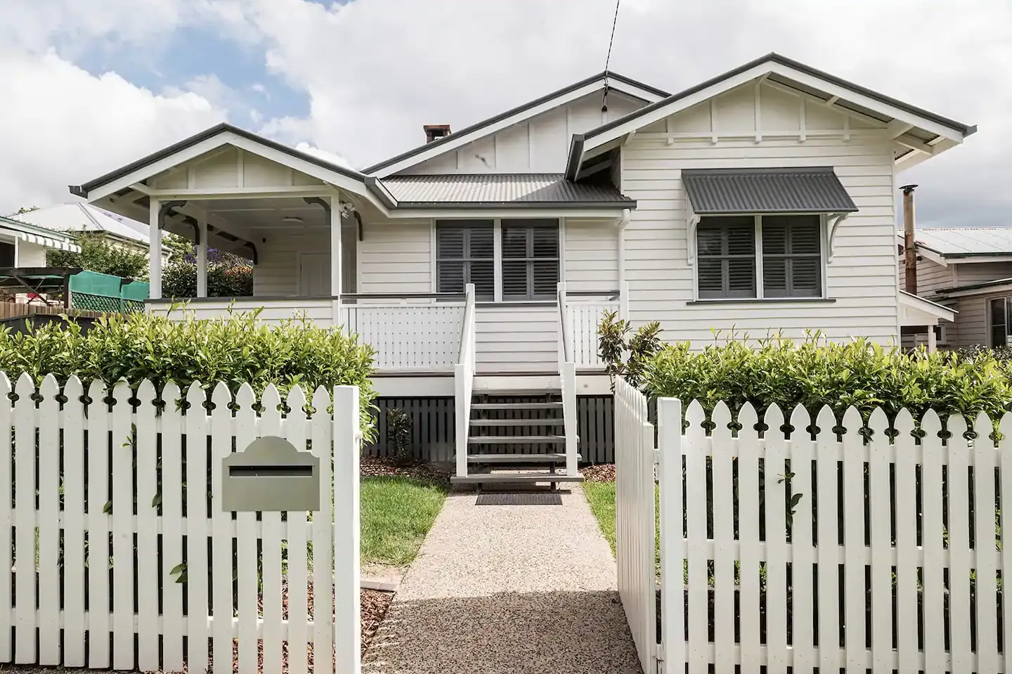 A charming white house with a gray roof and shutters sits behind a white picket fence. A concrete path leads through a gate and up to the front porch, which features stairs and railings. Greenery surrounds the property under a partly cloudy sky.