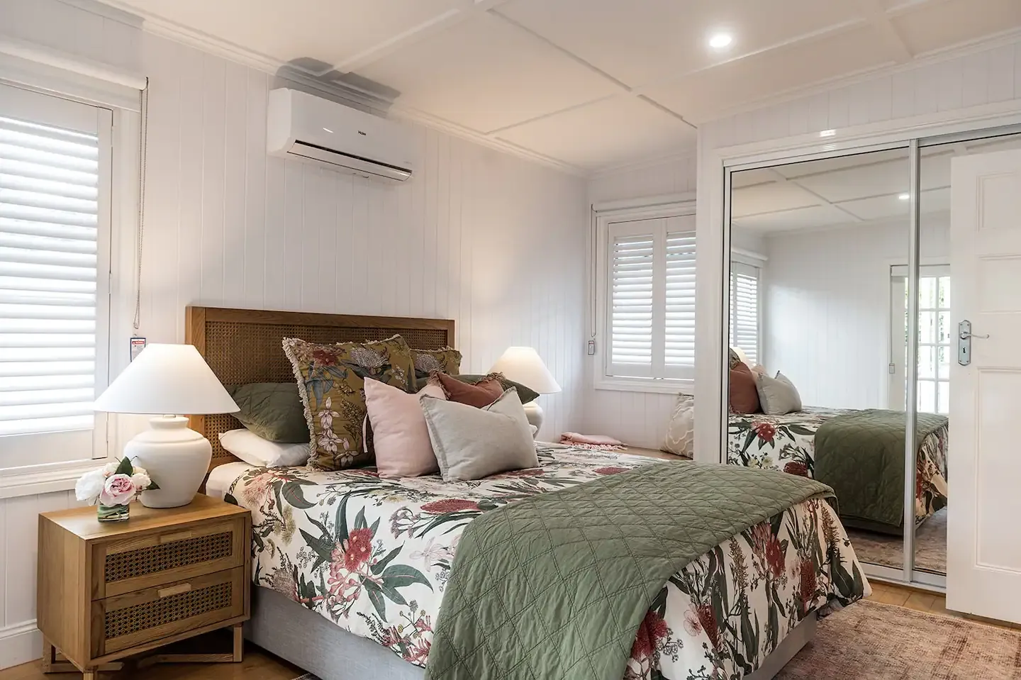 A cozy bedroom featuring a bed with floral and leafy patterned bedding, green quilt, and assorted pillows. A nightstand with a lamp and decorative items stands beside the bed. Large windows with shutters and a mirrored sliding closet door enhance the room's spacious feel.