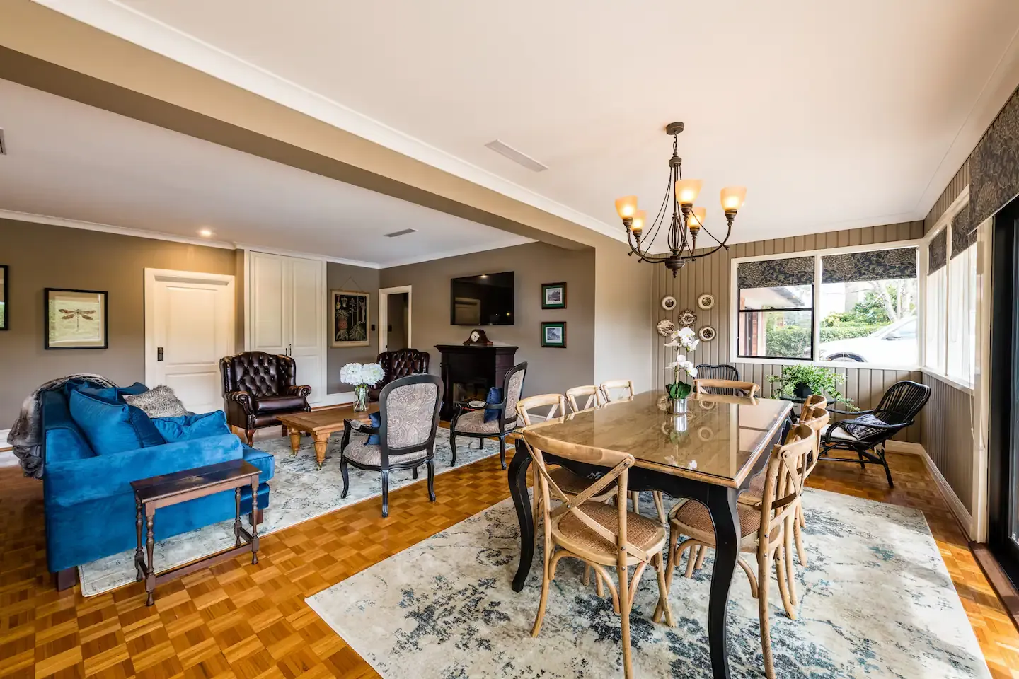 A cozy, well-lit living and dining room with parquet flooring. The space features a blue couch, two leather armchairs, a TV, and a fireplace. The dining area includes a table with eight chairs, a chandelier, and large windows providing ample natural light.