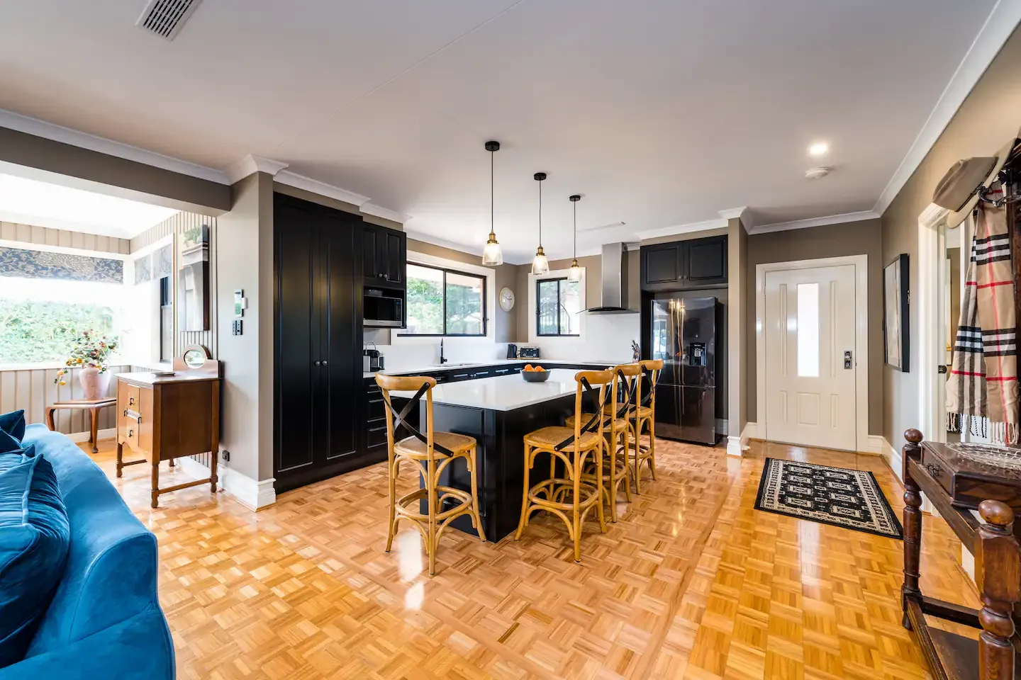 A modern kitchen with black cabinets, a large island with a black countertop and six wooden high chairs. Pendant lights hang above the island. The space features light wood parquet floors and adjacent seating area with a blue sofa. A front door is visible.
