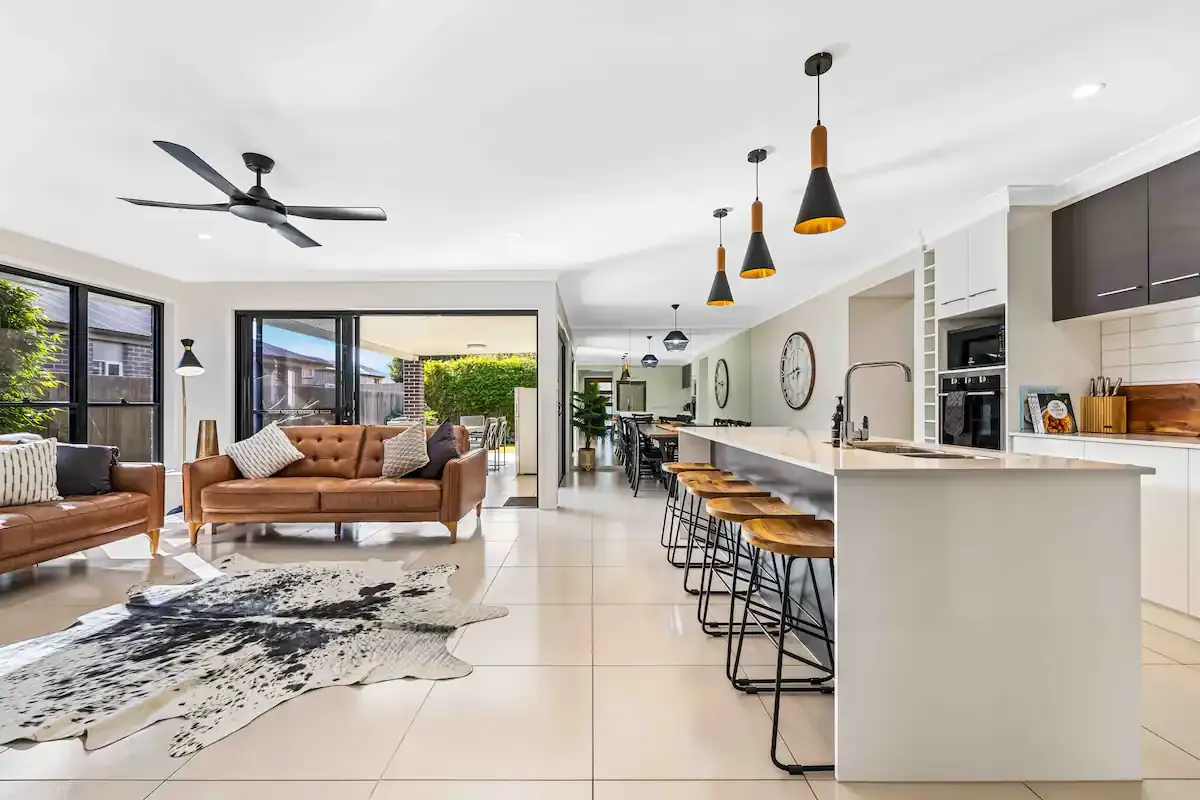 Modern open space interior with a kitchen and living area. The kitchen features a long island with bar stools, pendant lighting, and contemporary appliances. The living area has a brown leather sofa, a cowhide rug, and large glass doors opening to the patio.