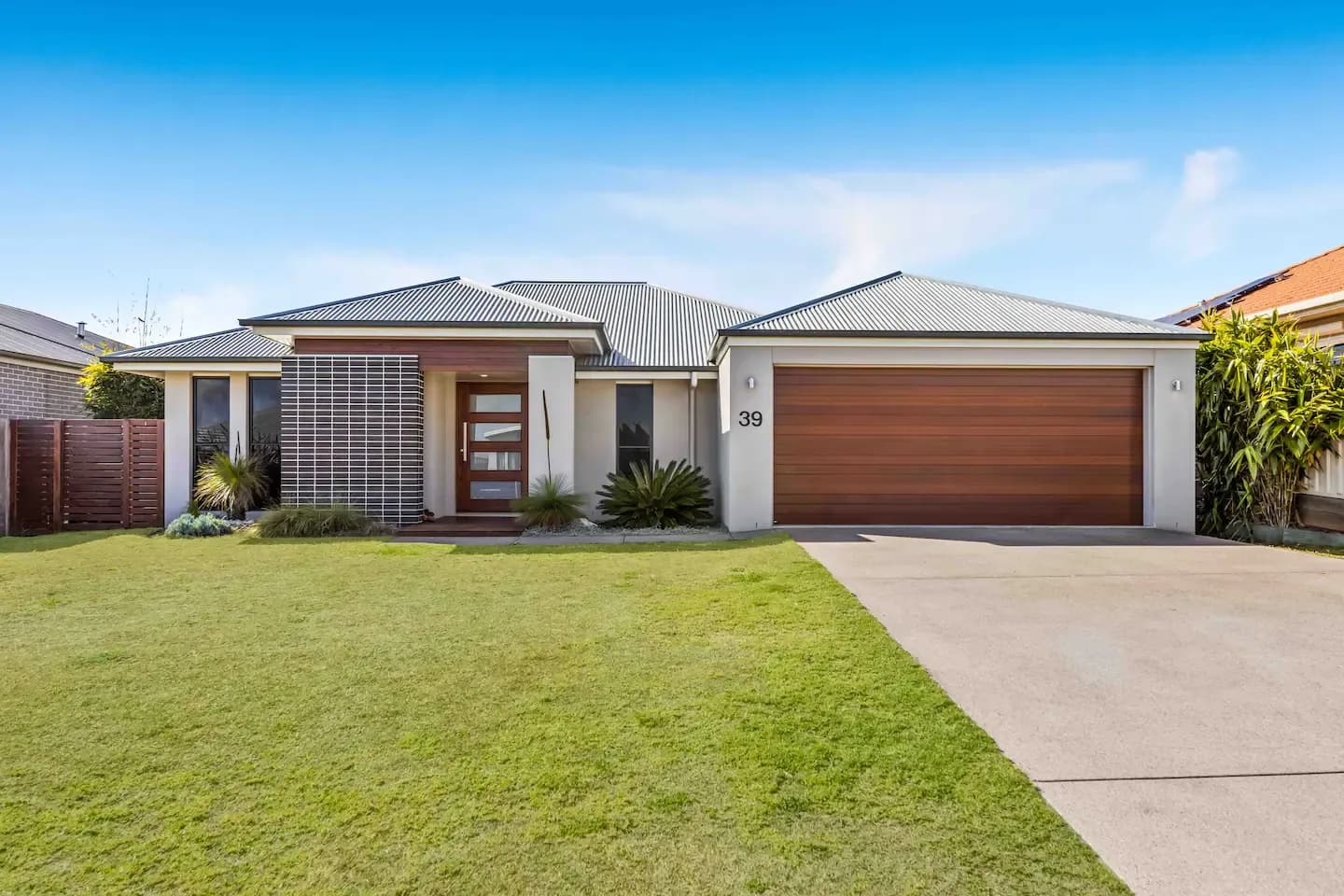 A modern, single-story house with a green lawn, two-car garage with wooden doors, and an inviting main entrance featuring a wooden door and side panel with decorative horizontal slats. The house has a minimalist, contemporary design and is surrounded by neatly trimmed plants.