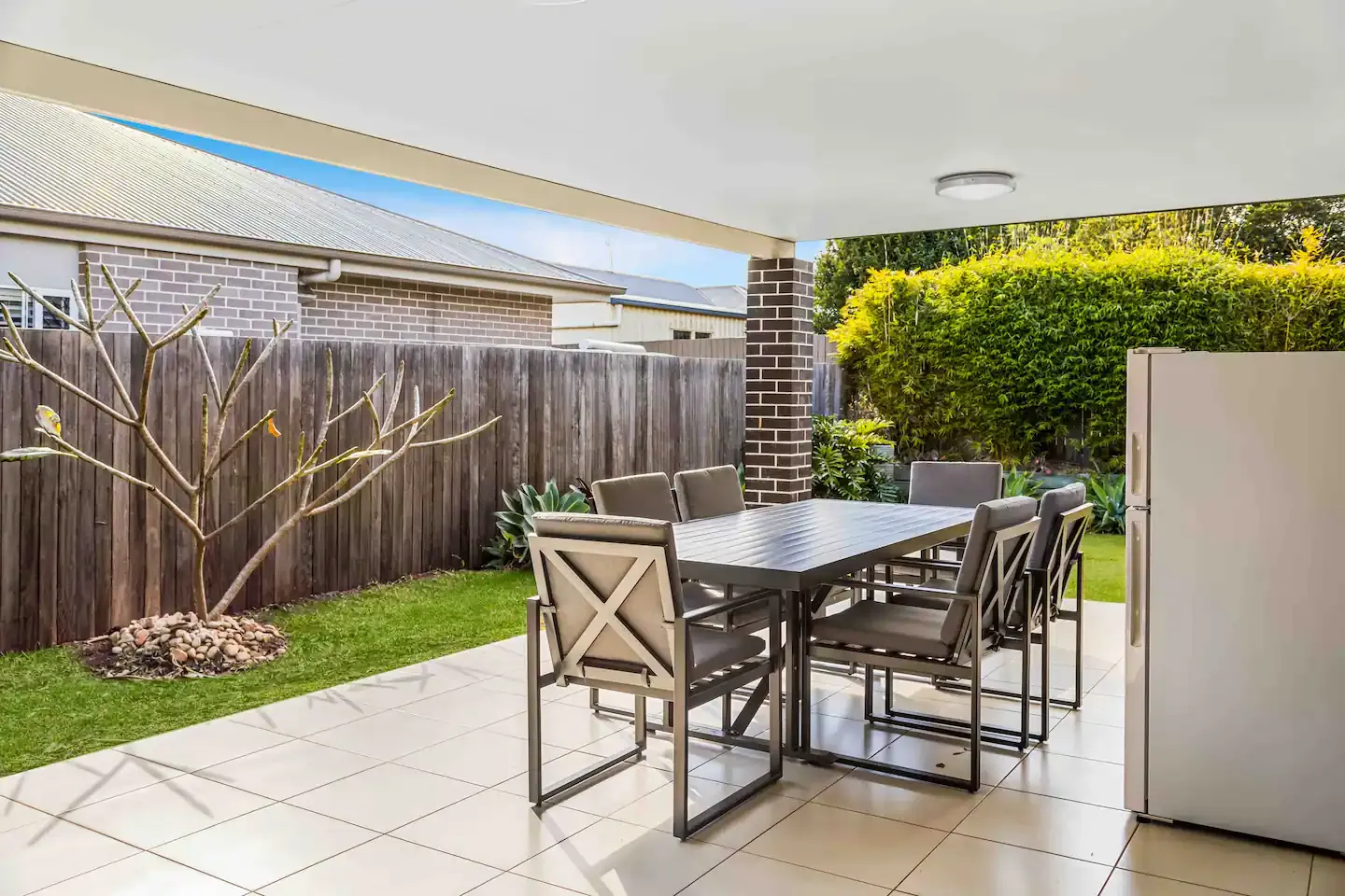 A covered outdoor patio with a dining table and six chairs on a tiled floor. The patio is adjacent to a grassy yard with a small tree and plants, enclosed by a wooden fence. A brick pillar and part of a house are visible in the background.