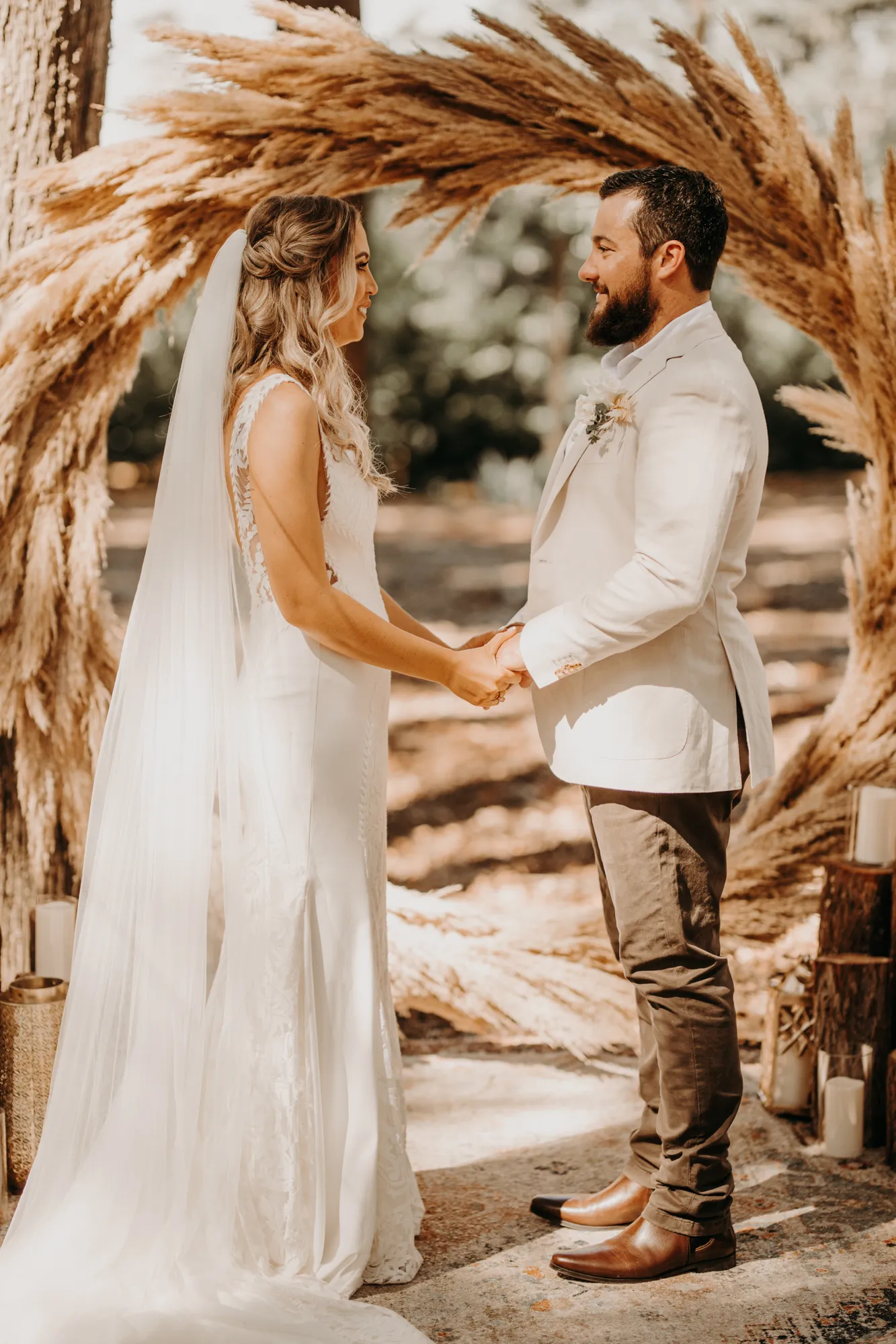 A bride and groom stand facing each other and holding hands outdoors at their wedding. The bride wears a white dress with a long veil, while the groom dons a white jacket and black pants. They are surrounded by a large, rustic, circular pampas grass arch.