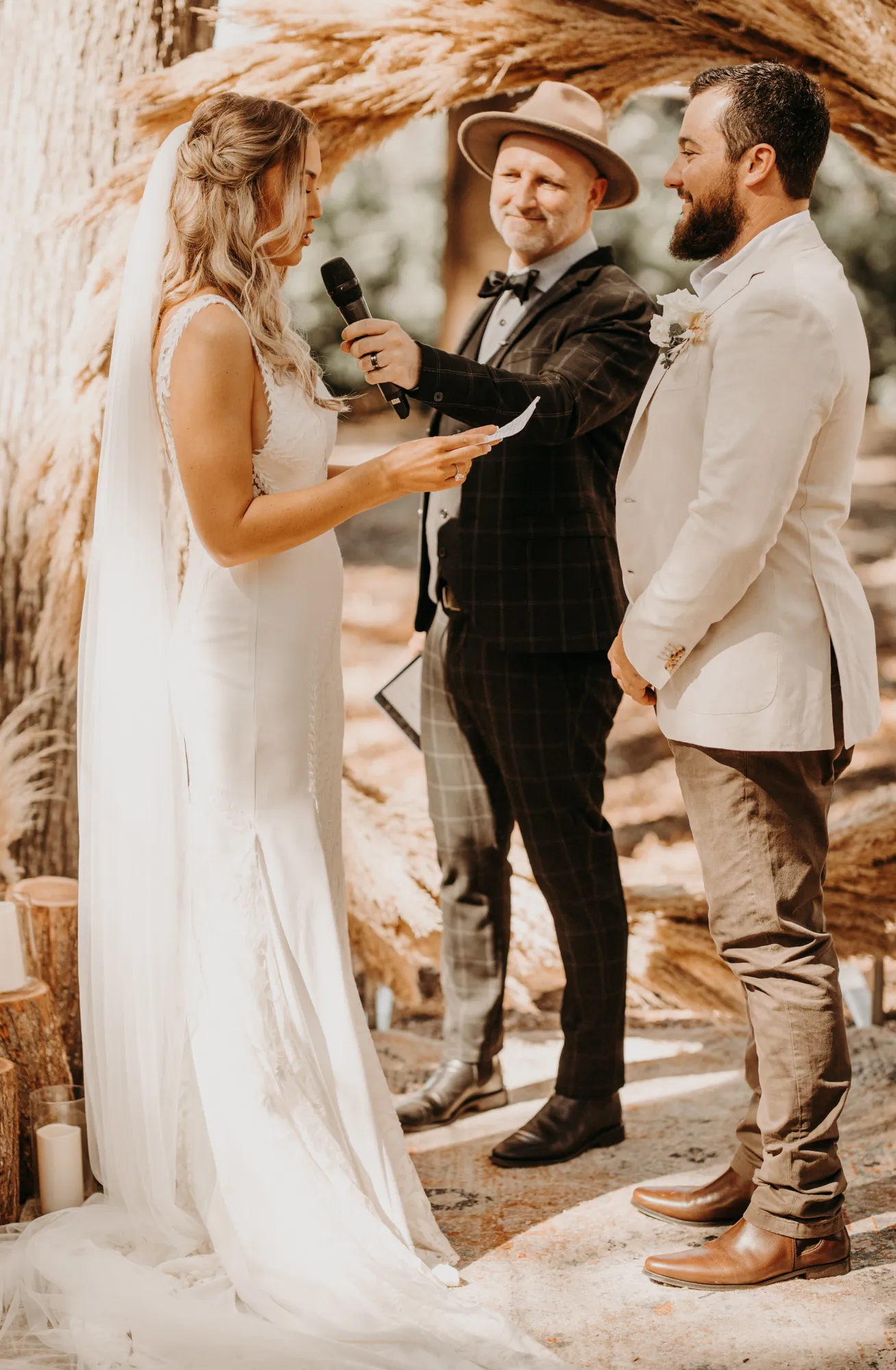 A couple stands facing each other during their outdoor wedding ceremony. The bride, in a white gown and veil, reads from a piece of paper, while the groom, in a white jacket and brown slacks, smiles at her. The officiant, in a plaid suit and hat, holds a microphone.