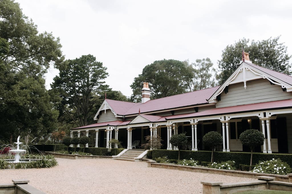 A historic-style, single-story house with a red roof, white accents, and multiple gables, set amid lush greenery. A gravel path in the foreground leads to the entrance, flanked by manicured shrubs. A fountain is visible to the left, enhancing the picturesque scene.