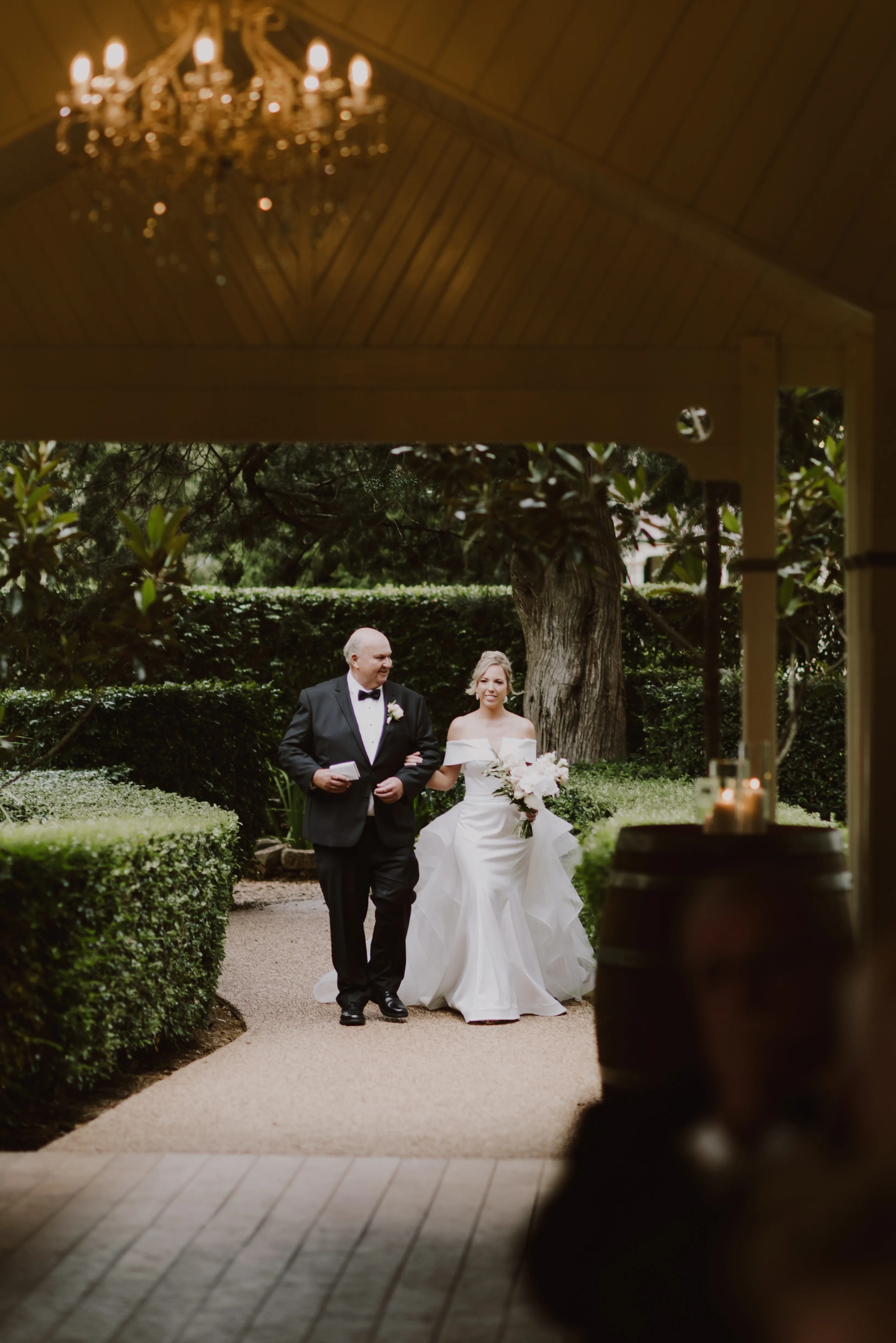 A bride in a white dress is walking arm-in-arm with an older man in a black suit and bow tie. They are on a garden pathway framed by lush greenery, approaching an indoor area lit by a chandelier. The bride is holding a bouquet of white flowers.