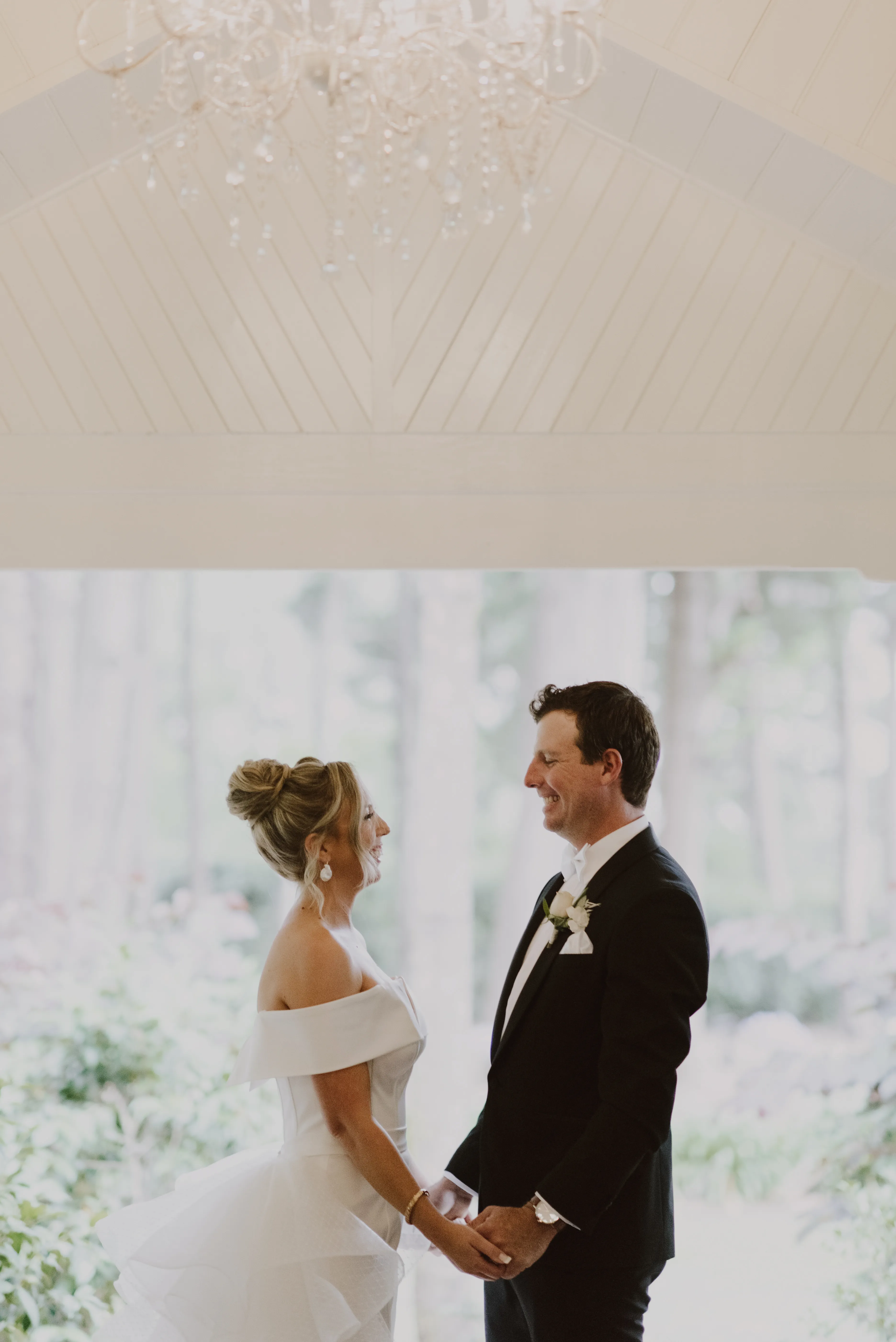 A bride and groom stand facing each other while holding hands in a bright, airy room. The bride wears an off-the-shoulder wedding gown and the groom wears a black suit with a white boutonnière. They are smiling under a delicate chandelier.