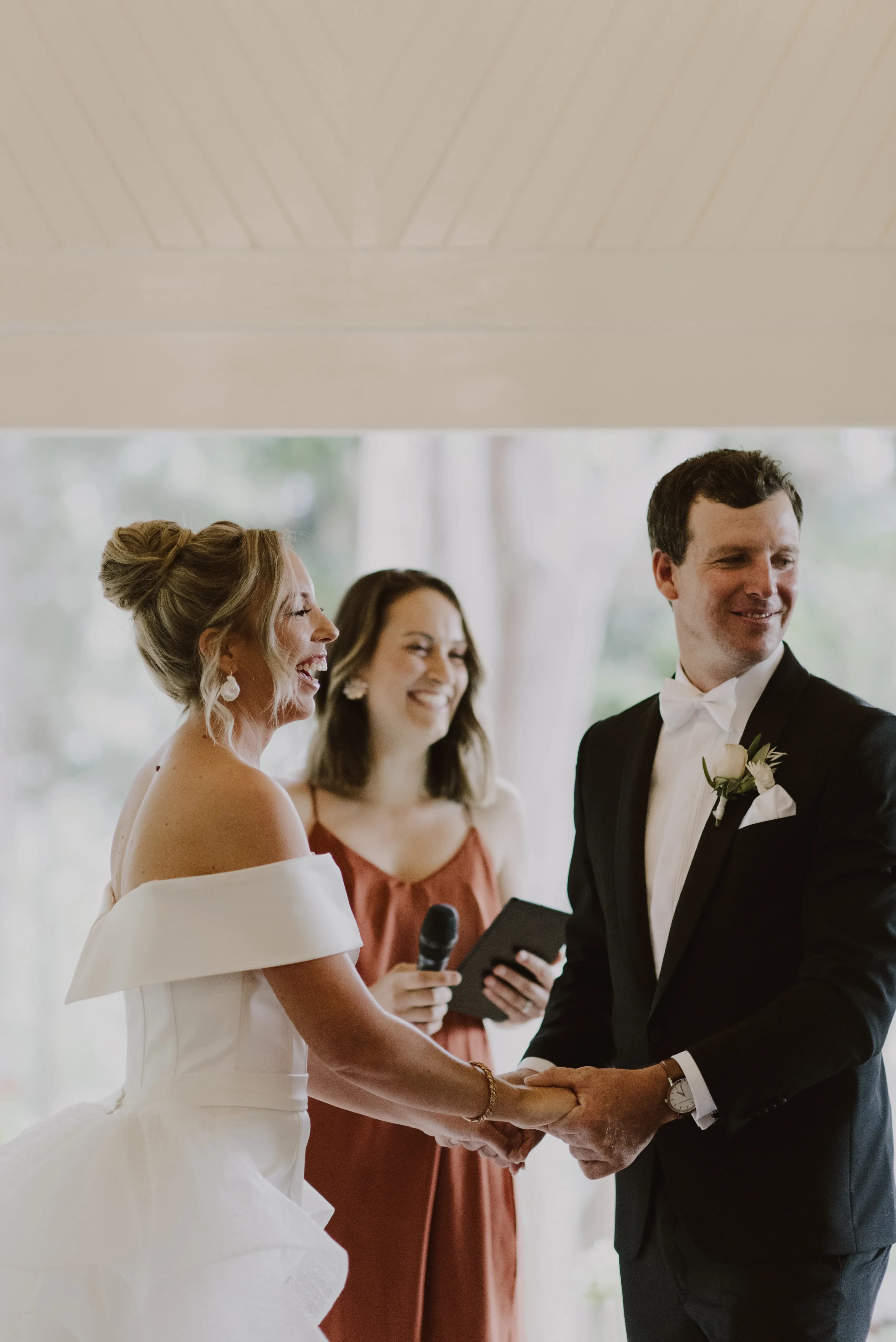A bride and groom laughing and holding hands during their wedding ceremony. The bride is wearing an off-the-shoulder white wedding dress, and the groom is in a black tuxedo with a white bow tie. A woman in a rust-colored dress stands behind them, holding a microphone and tablet.