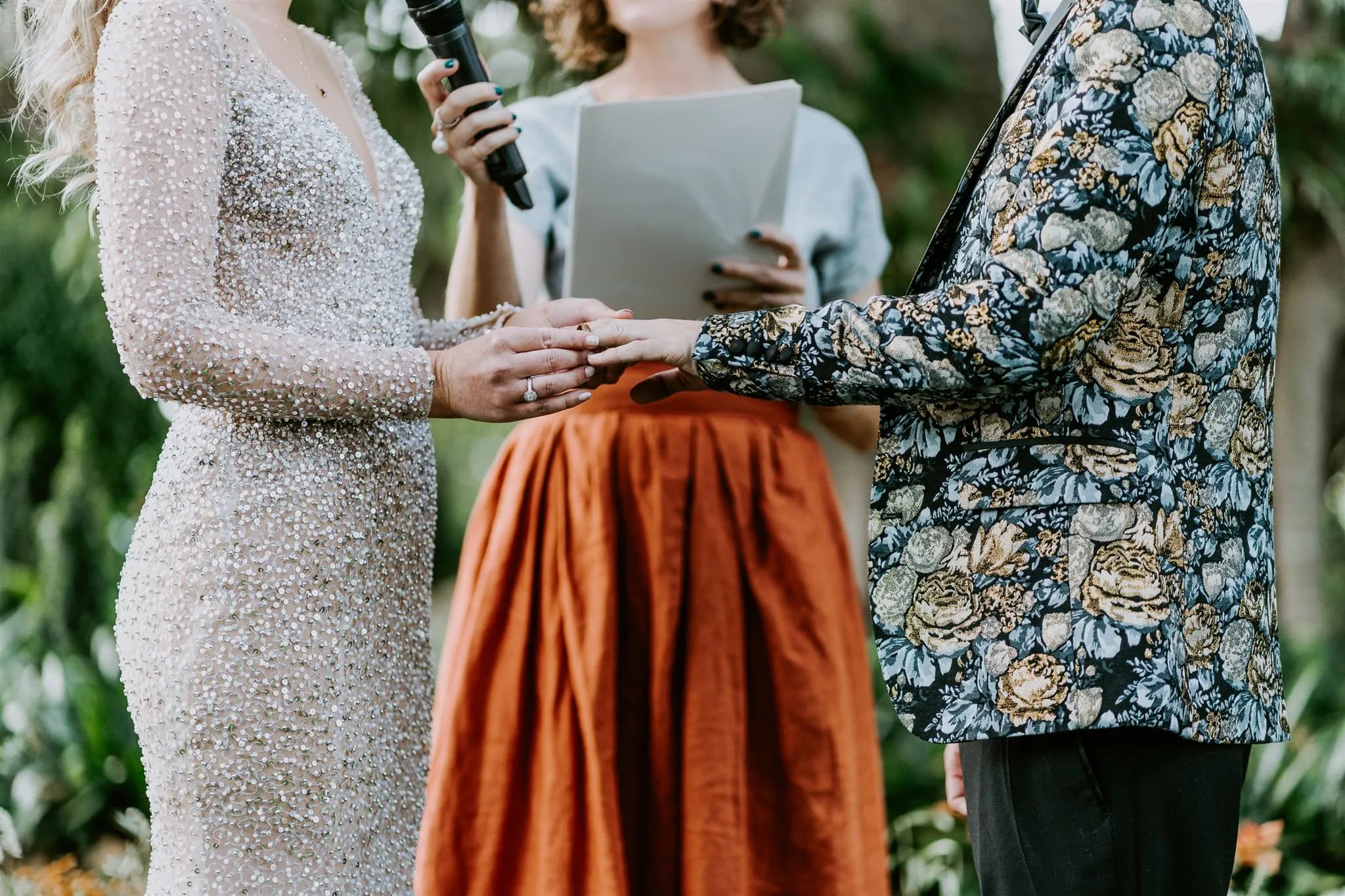 A couple is exchanging rings during their wedding ceremony. The bride is wearing a beaded, long-sleeved dress, while the groom is in a floral-patterned jacket. An officiant, holding a microphone and papers, stands between them. They are outdoors, surrounded by greenery.