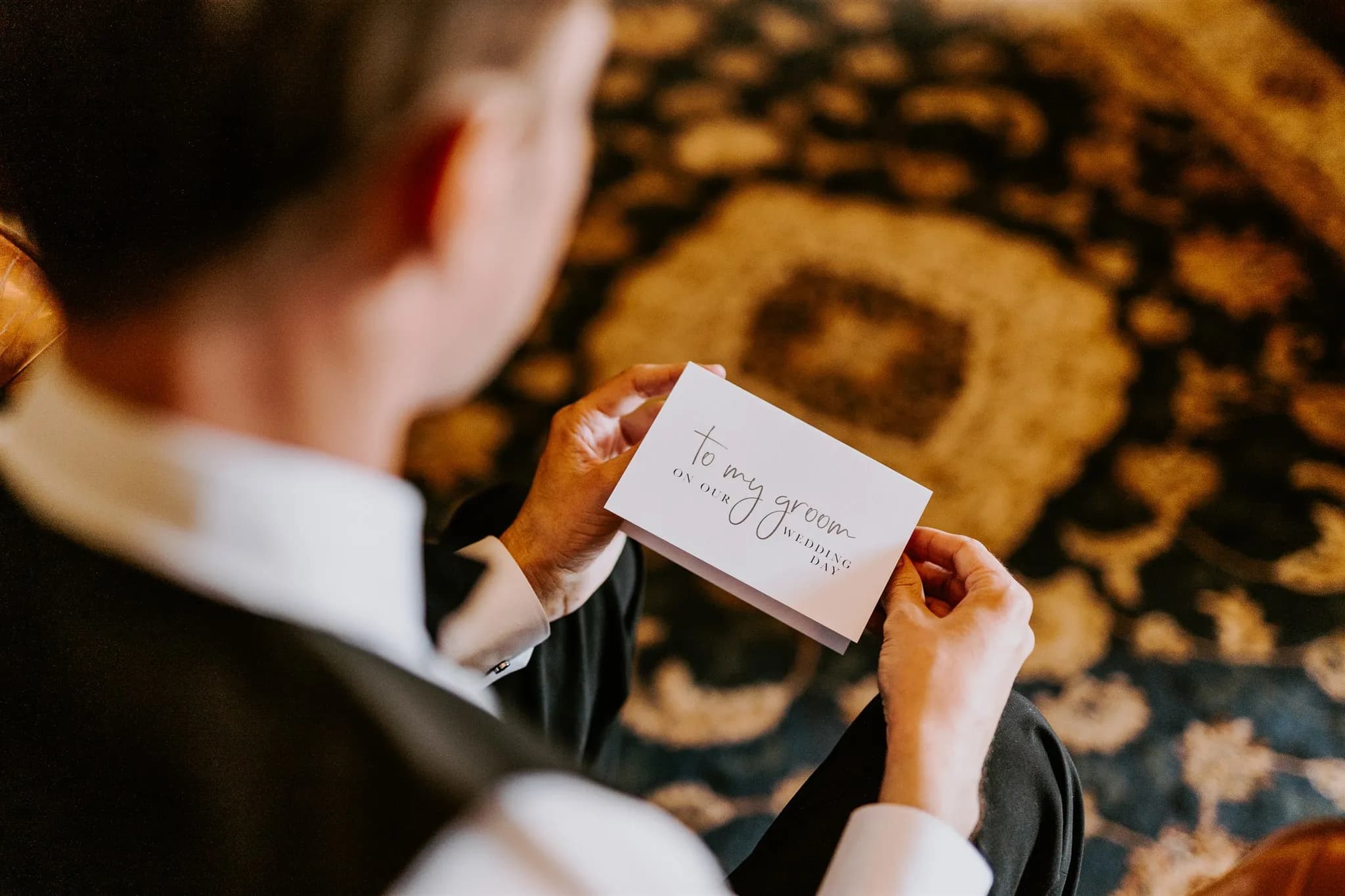 A person in formal attire is holding an envelope with the handwritten text "To my groom on our wedding day." The person is seated, and a patterned carpet is visible in the background. The image focuses on the envelope and the hands holding it.