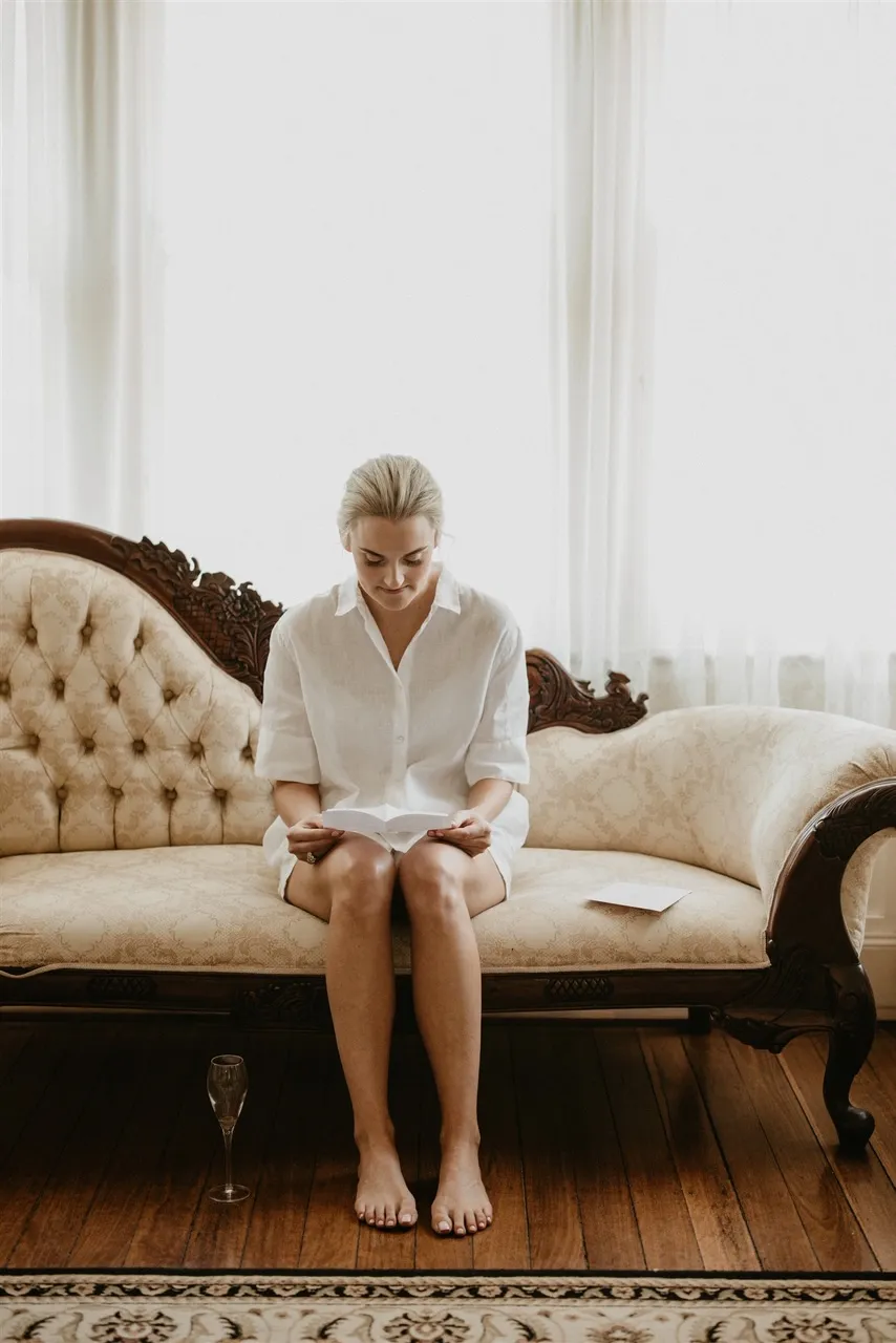 A woman in a white dress sits on a vintage-style, cream-colored, tufted couch in a sunlit room with wooden floors. She is reading a letter or card. A glass with a stem, possibly for wine or champagne, is placed on the floor in front of her.