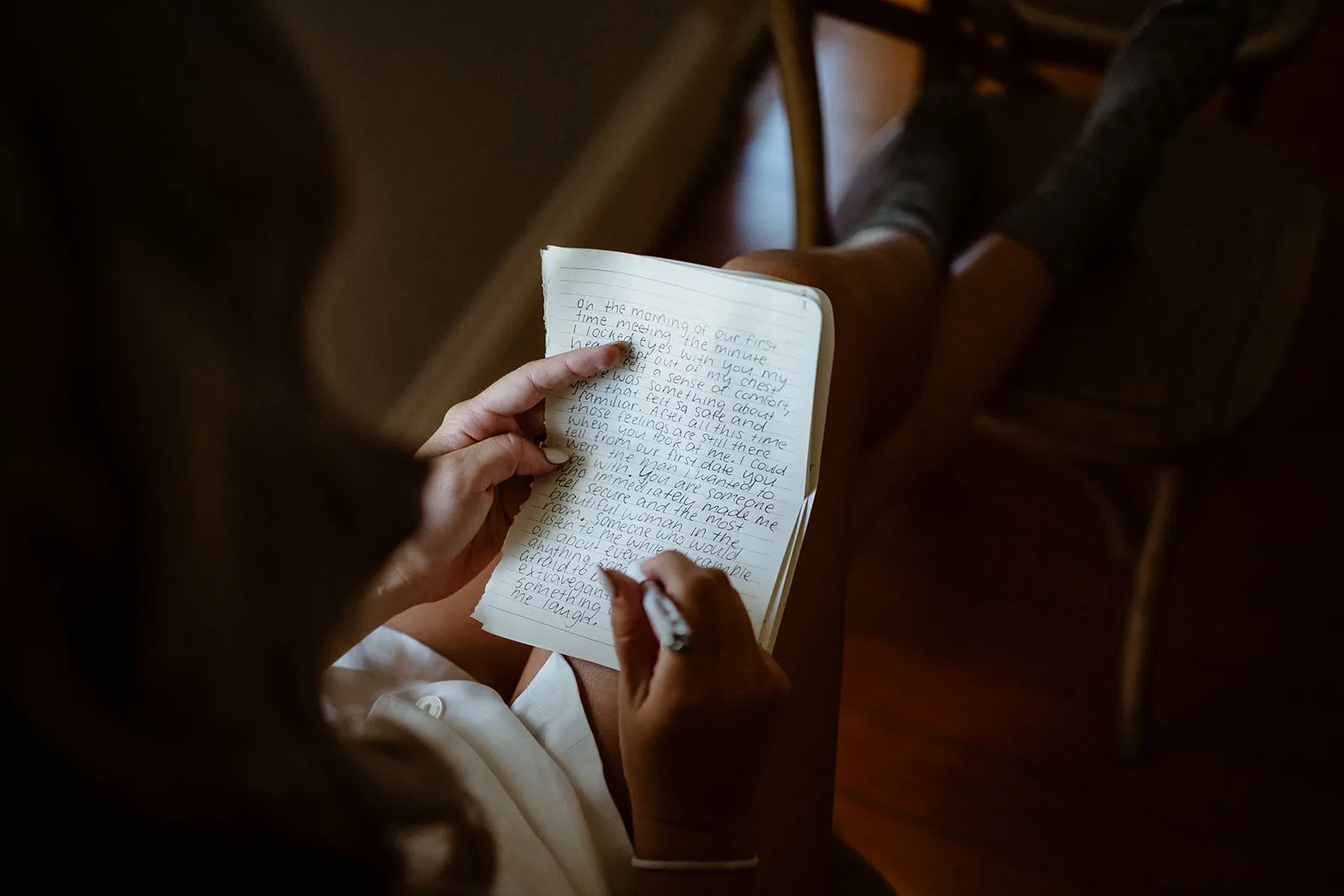 Person sitting and writing in a notebook, with their hand pointing at a line of text. The person is indoors, wearing socks, and their pen is poised over the notebook. The light casts a warm tone over the scene.