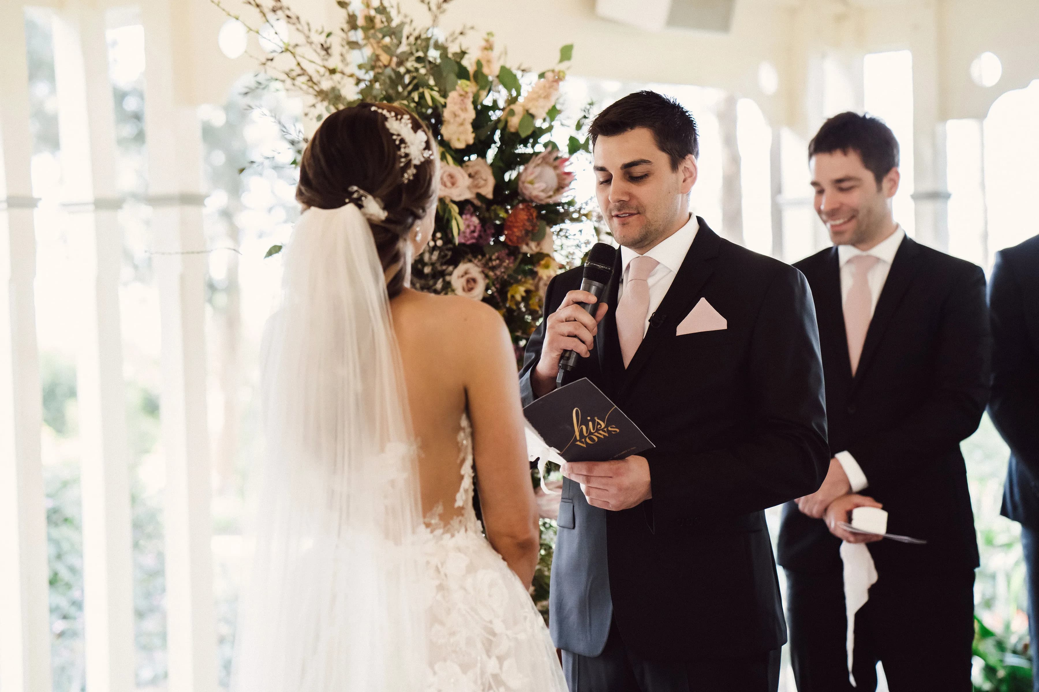 A bride and groom stand together during their wedding ceremony. The groom, dressed in a black suit with a pink tie and pocket square, reads from a card while holding a microphone. The bride, in a white gown with a veil, faces him. A man in a suit stands behind them, smiling.