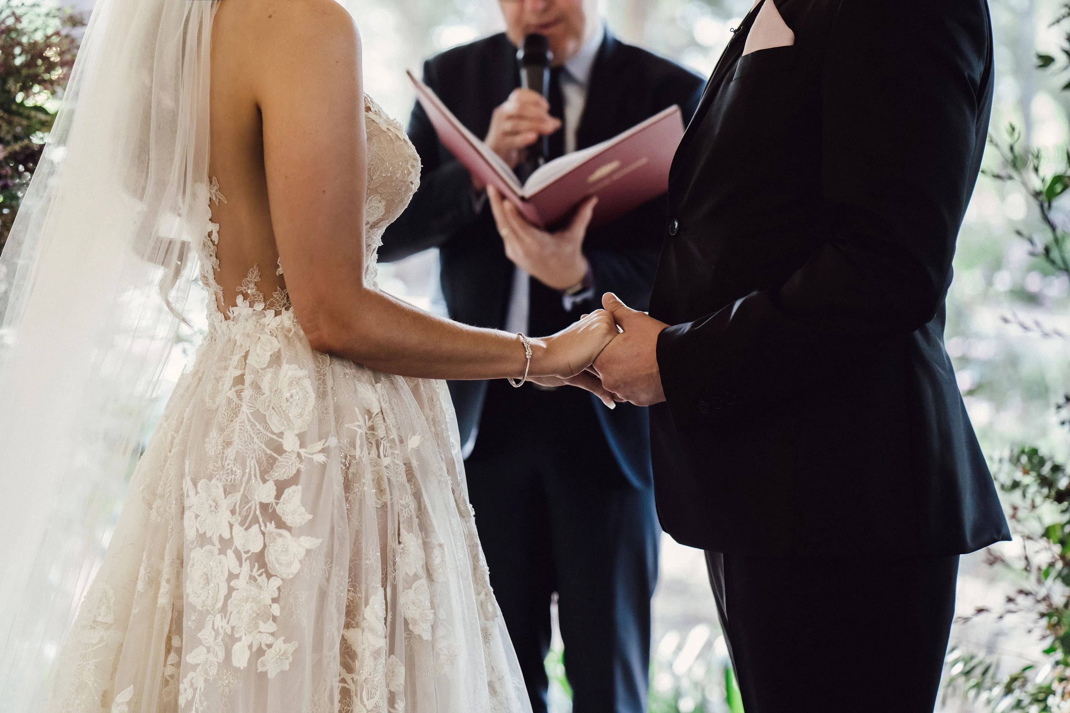A bride and groom hold hands during a wedding ceremony. The bride wears a lace wedding dress with a veil, and the groom wears a black suit. A person stands behind them holding a microphone and a book, possibly officiating the ceremony.