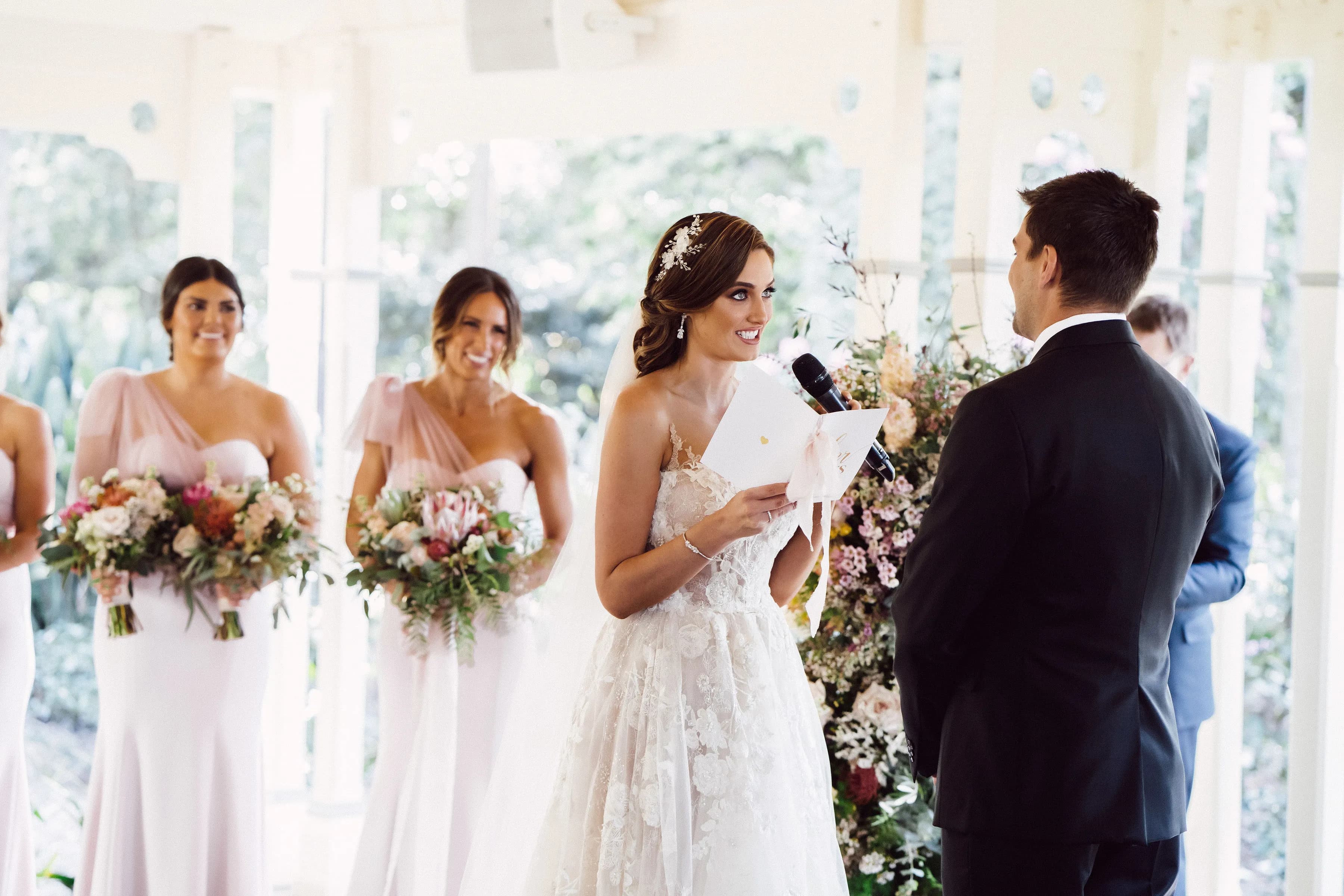 A bride, dressed in a floral lace gown, reads her vows to her groom, who is wearing a black suit. She holds a microphone and a card. Three bridesmaids stand beside her, holding bouquets of flowers and dressed in pale pink dresses. The scene is set in an outdoor venue.