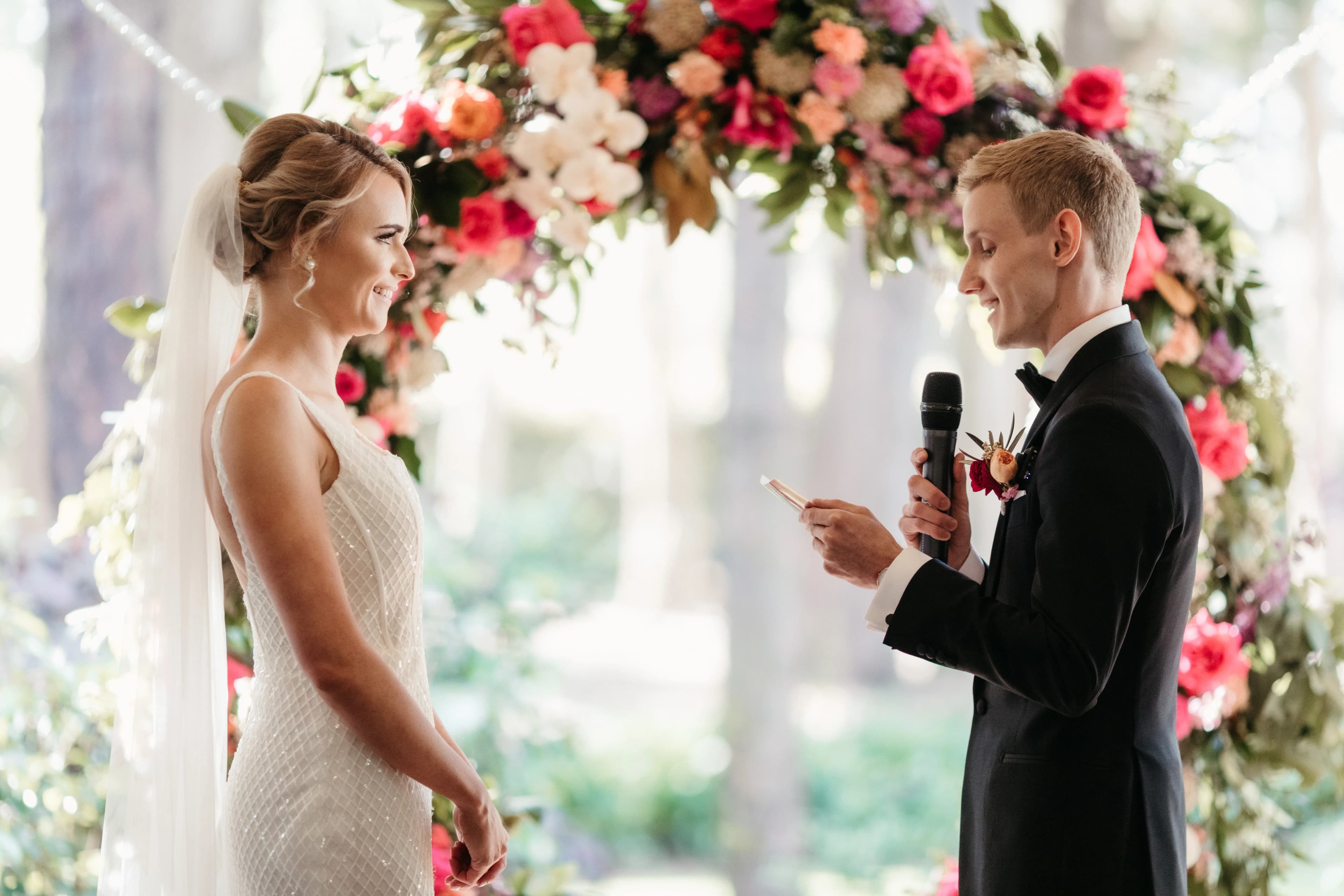 A bride in a white dress and veil smiles at a groom in a black suit as he speaks into a microphone. They stand in front of a flower arch adorned with vibrant pink, red, and white flowers, with a forested background softly blurred behind them.