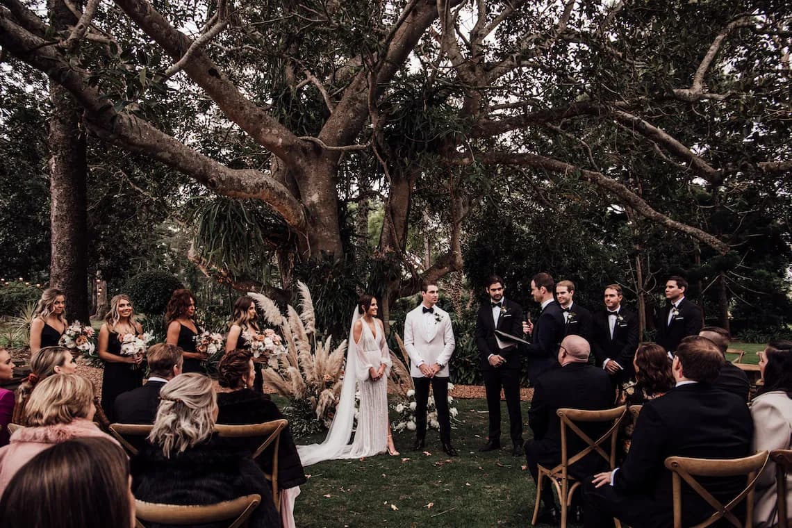 A couple stands under a large tree during their outdoor wedding ceremony. The bride, in a white gown, and the groom, in a white jacket and black pants, are surrounded by their bridal party. Guests seated on wooden chairs watch attentively.