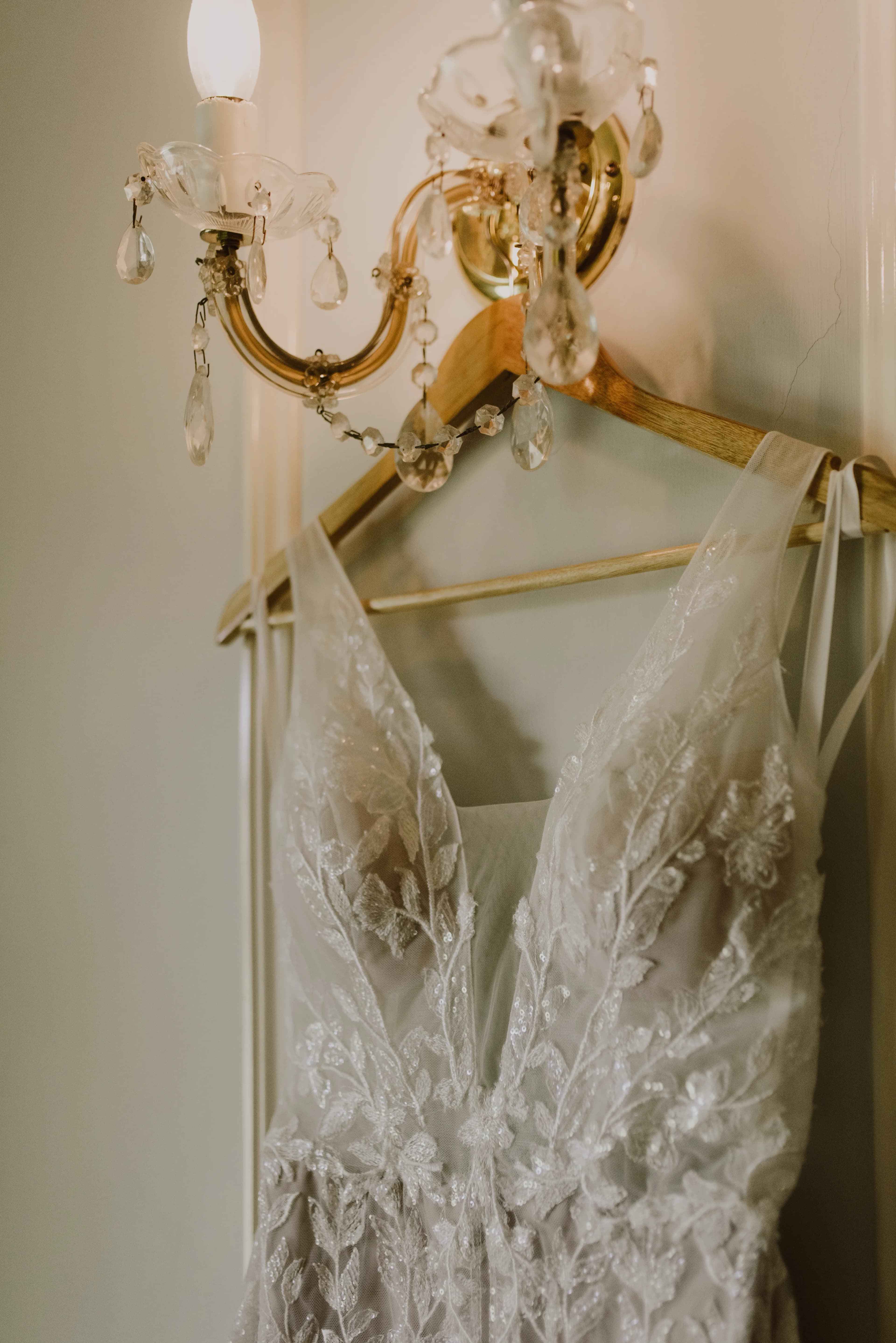 A detailed white lace bridal gown hangs from a wooden hanger on a brass sconce adorned with dangling crystals. The intricate patterns and sheer fabric of the dress are illuminated softly by the light from the sconce, creating an elegant and delicate ambiance.